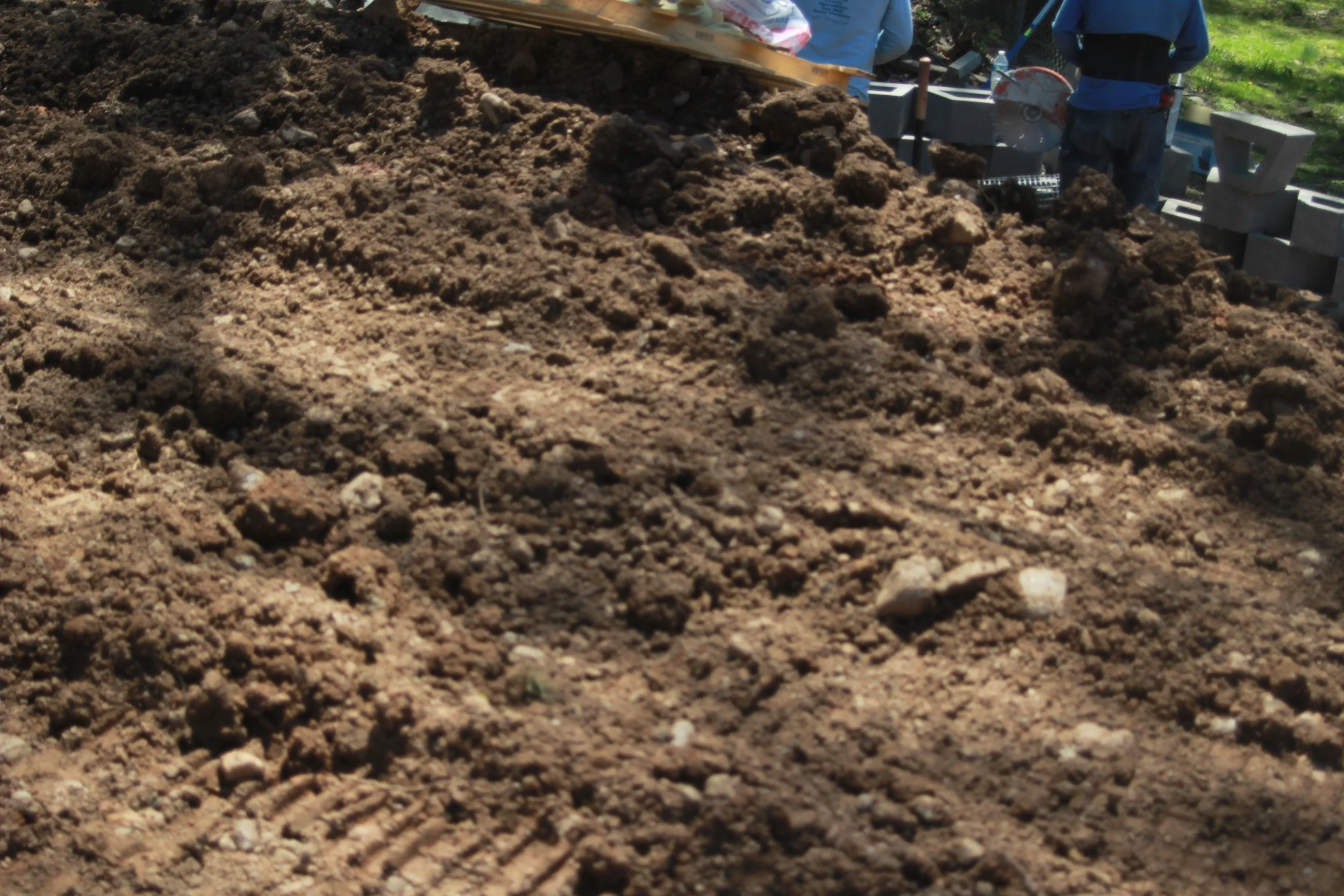 Close-up view of freshly dug soil with small rocks, with construction workers and concrete blocks in the background.