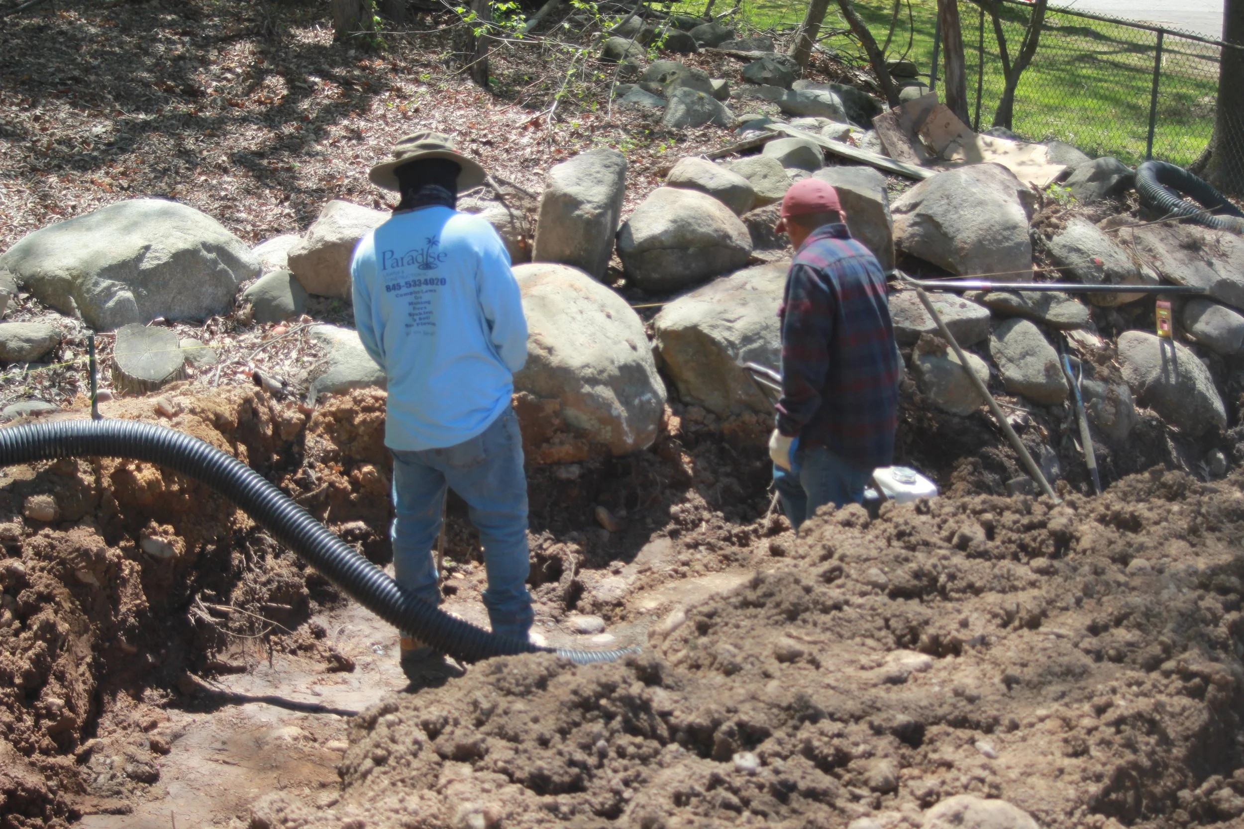 Two workers in casual clothing and hats are working outdoors on a construction or landscaping project, with rocks and soil scattered around, and some equipment visible.