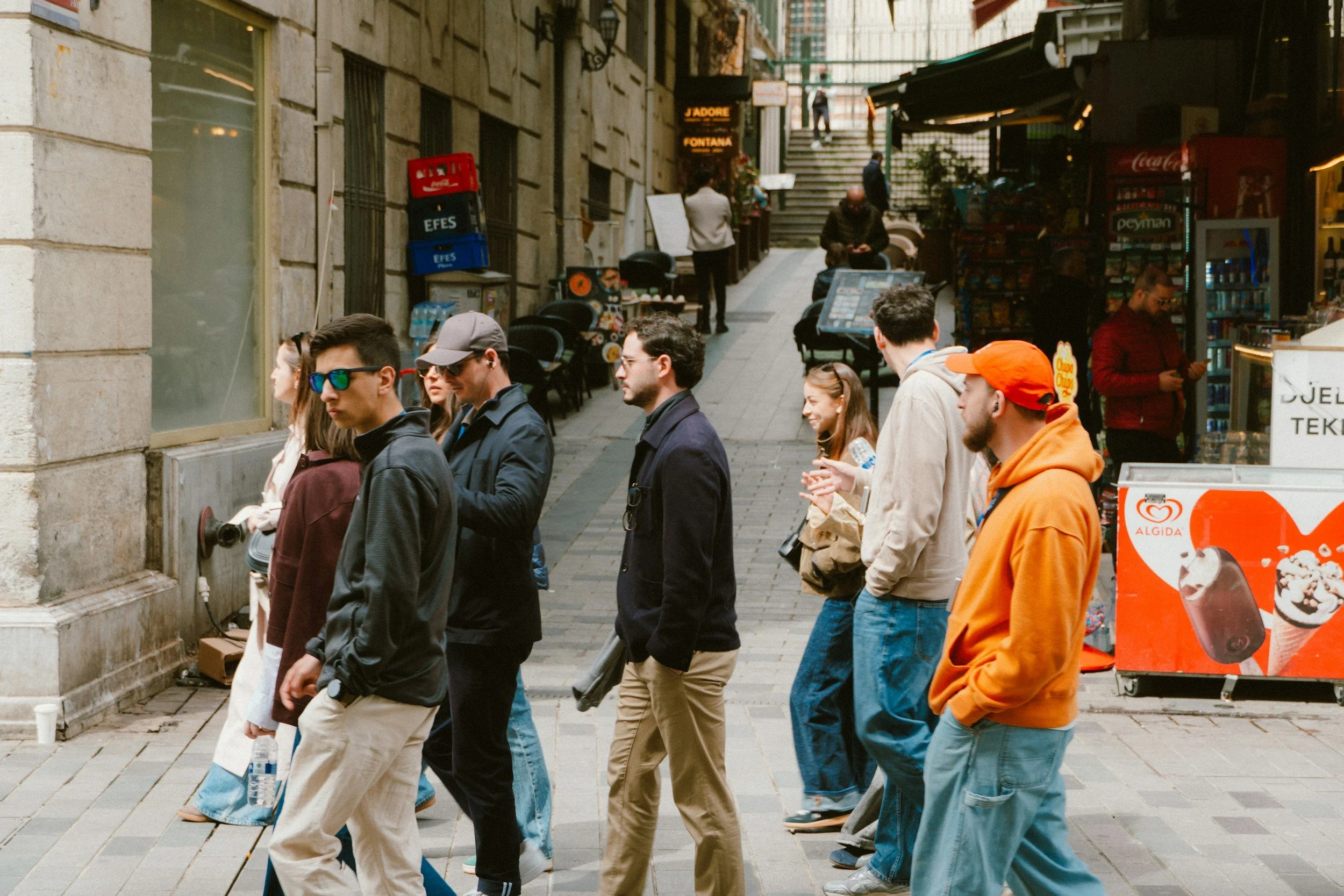 A group of people walking on a city street sidewalk with storefronts and stairs in the background.
