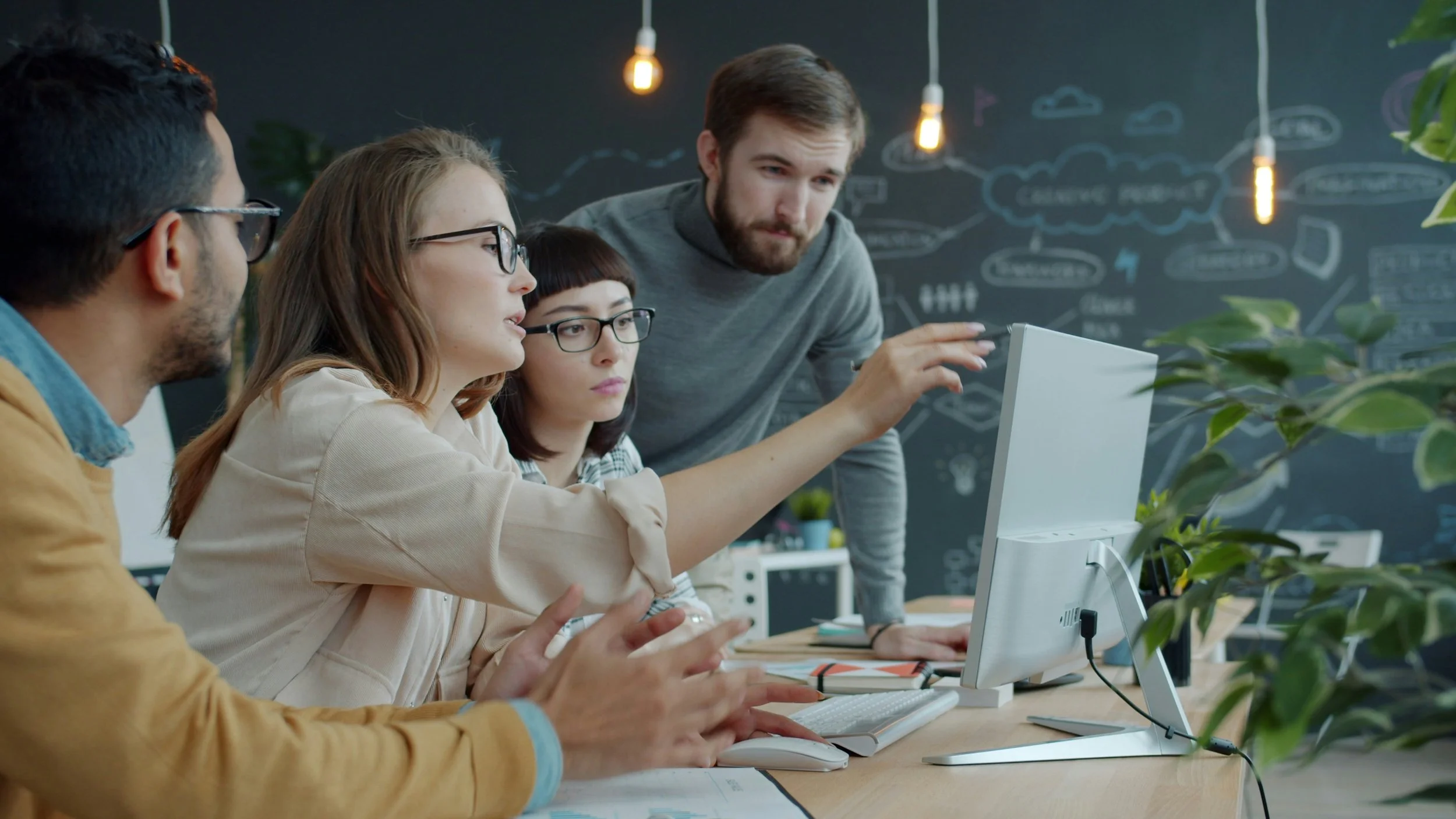 Group of four young adults working together at a computer in an office with a blackboard wall filled with drawings and notes in the background.