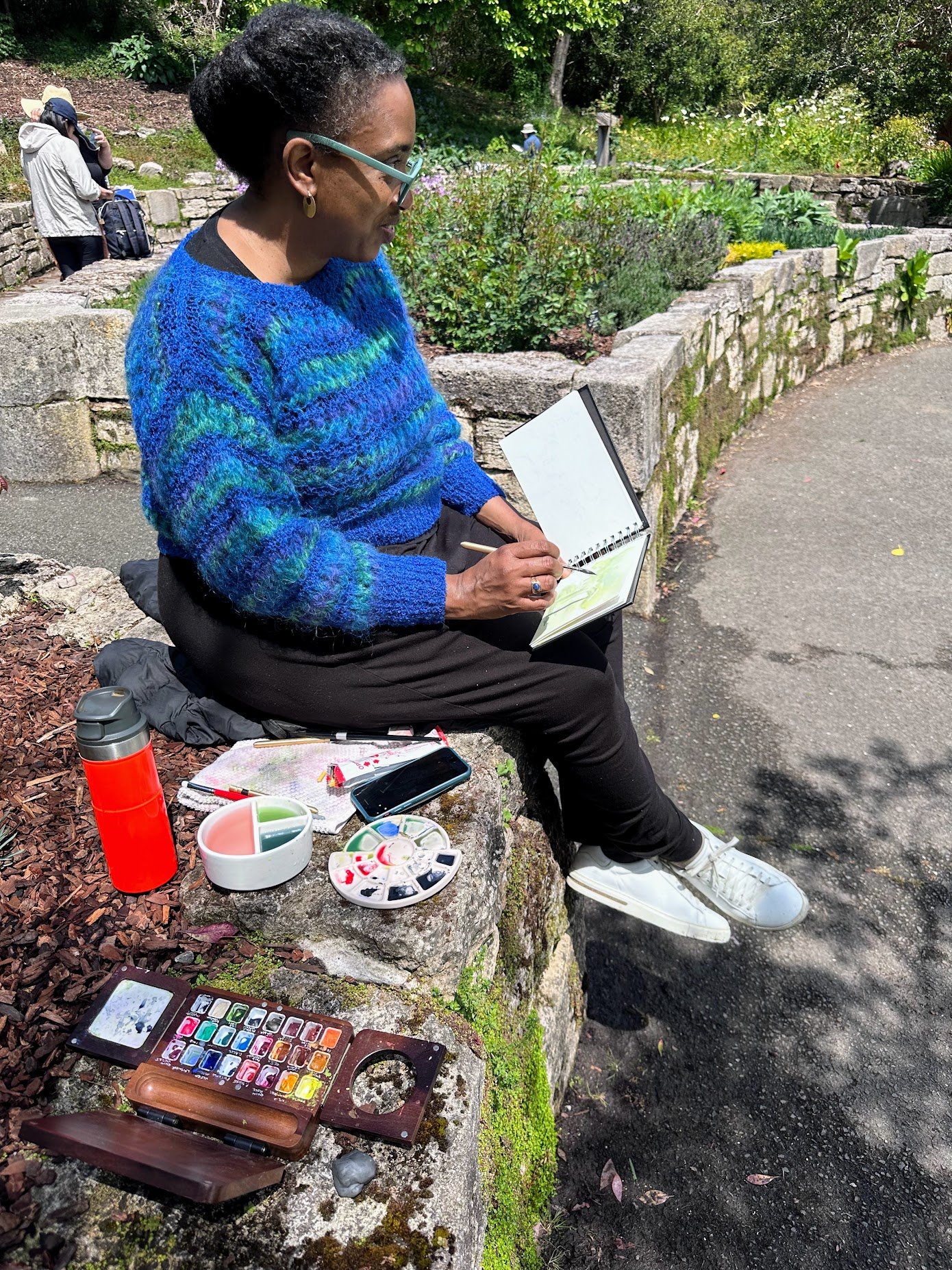 A woman sitting on a stone bench outdoors, painting in a sketchbook with watercolor paints and brushes nearby, surrounded by garden plants and other people in the background.