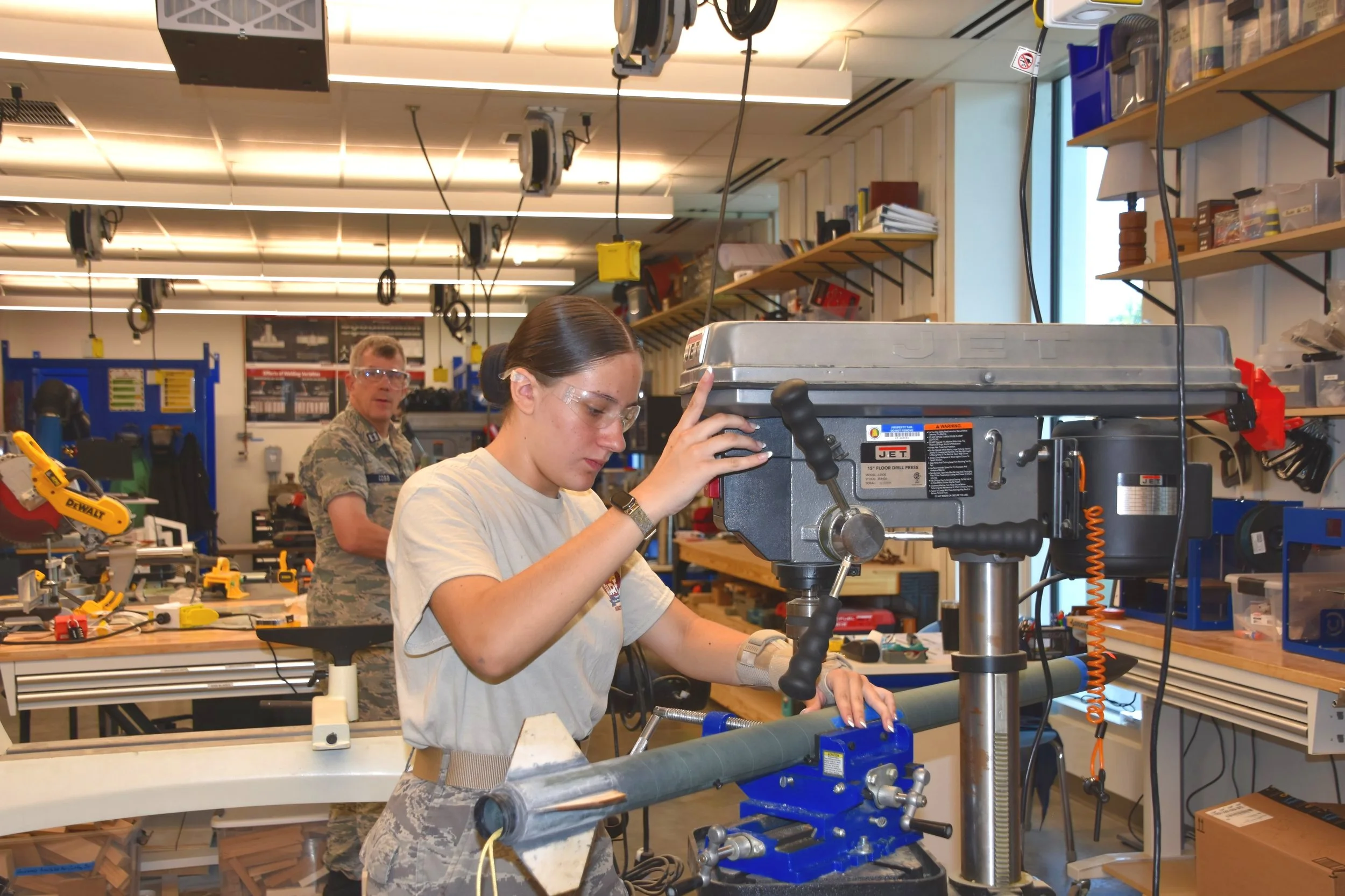 A young female cadet operating a drilling machine in a workshop, wearing protective eyewear in Civil Air Patrol's Aircraft Maintenance Academy. Another person in camouflage uniform is in the background. The workshop has various tools and equipment.