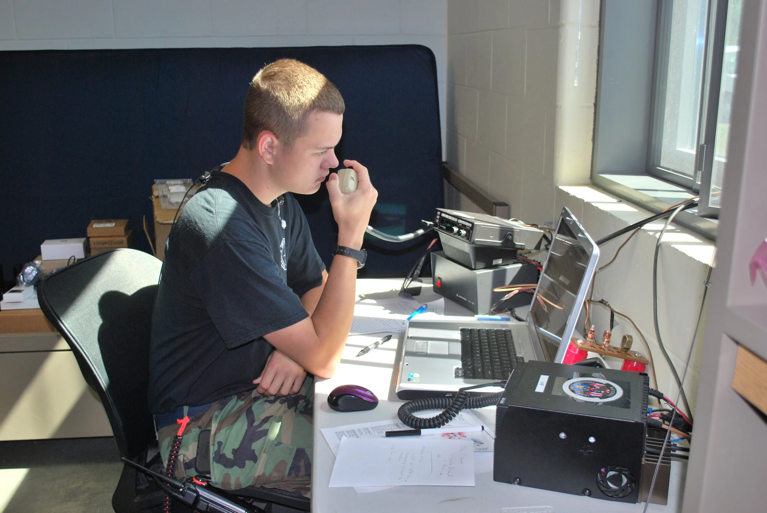 Person using a radio and laptop at a desk in an office setting.
