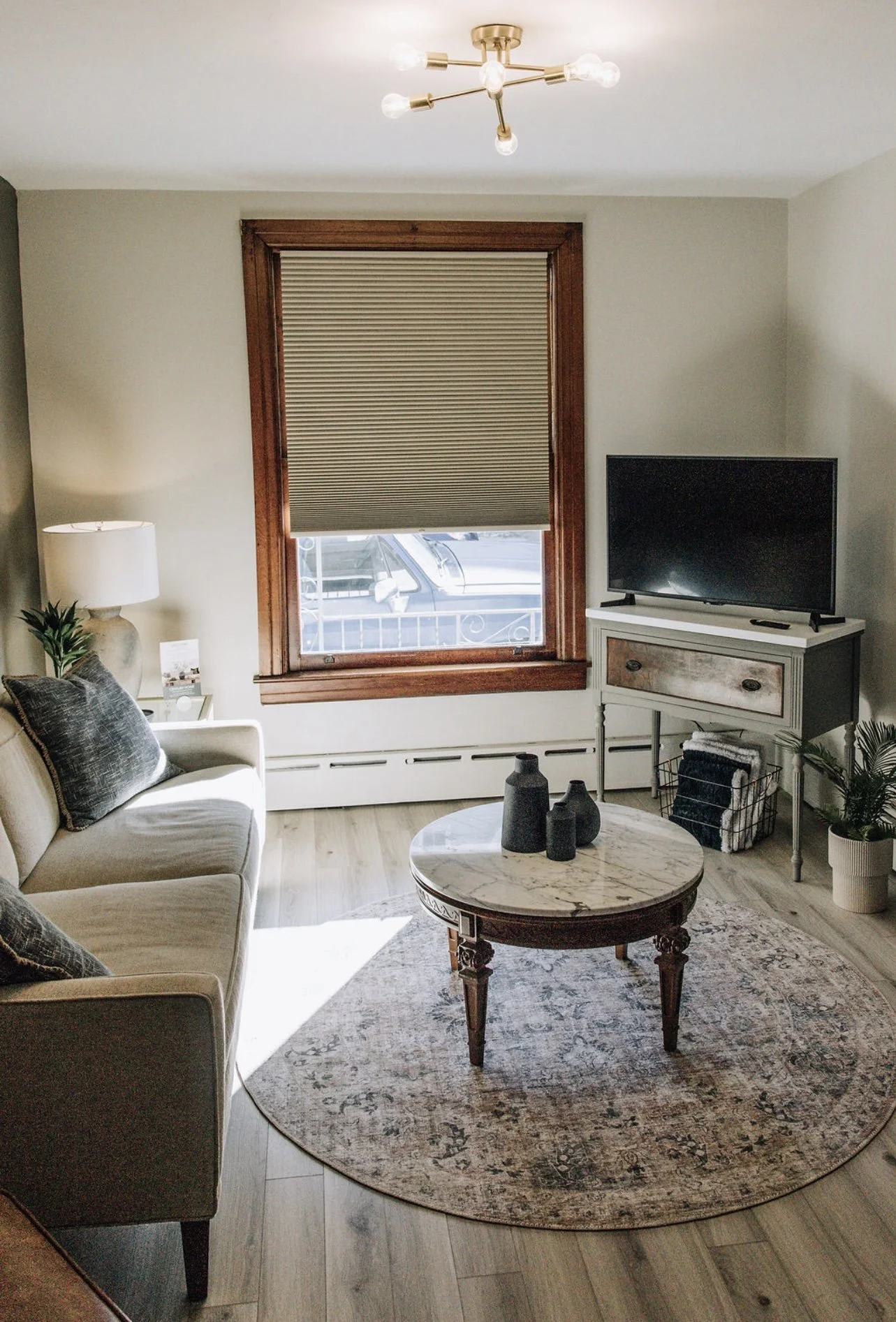 Living room with beige sofa, dark pillows, white lamp, round marble coffee table with black vases, patterned rug, window with wooden frame and window shade, TV on a white stand, and potted plant.