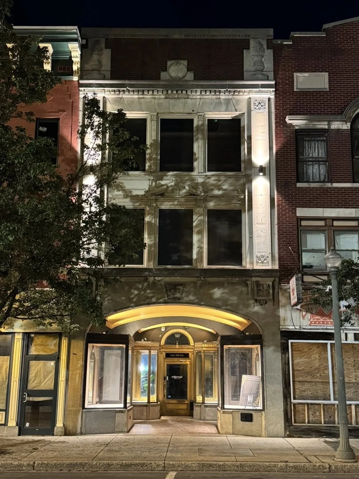 Nighttime view of a historic building with illuminated entrance, large windows, and decorative architectural details, flanked by neighboring brick buildings and a streetlamp.