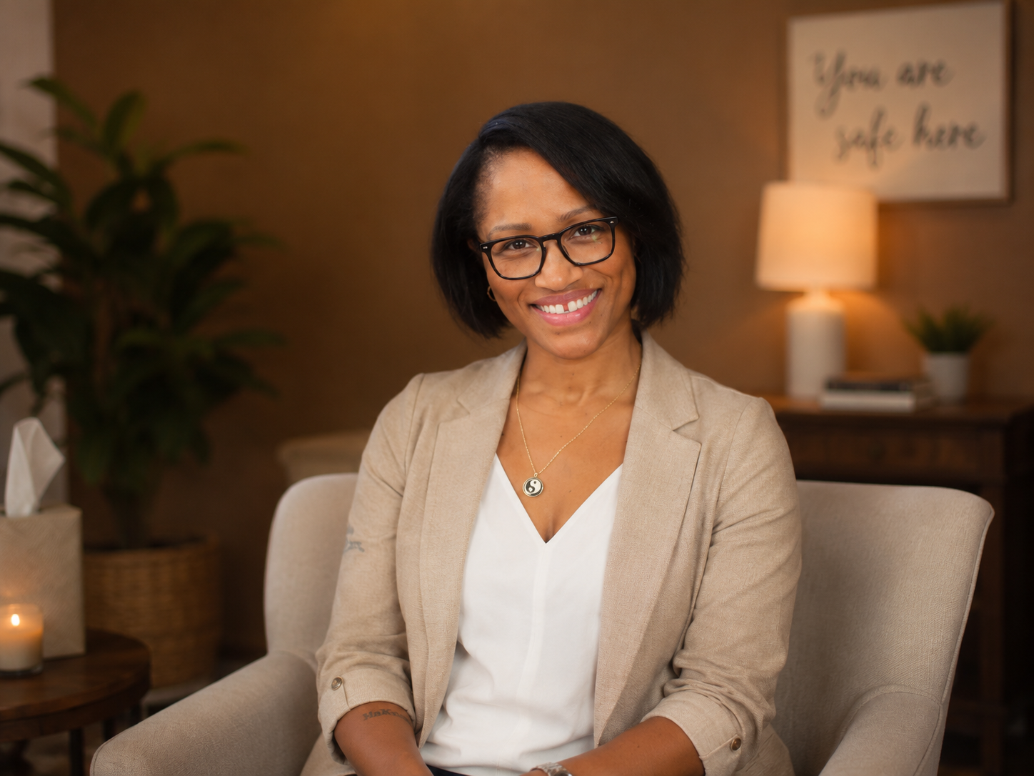 Smiling woman in glasses and beige blazer sitting on a beige chair in a cozy, warmly lit room with a lamp, a plant, and framed art that says 'You are safe here' in the background.