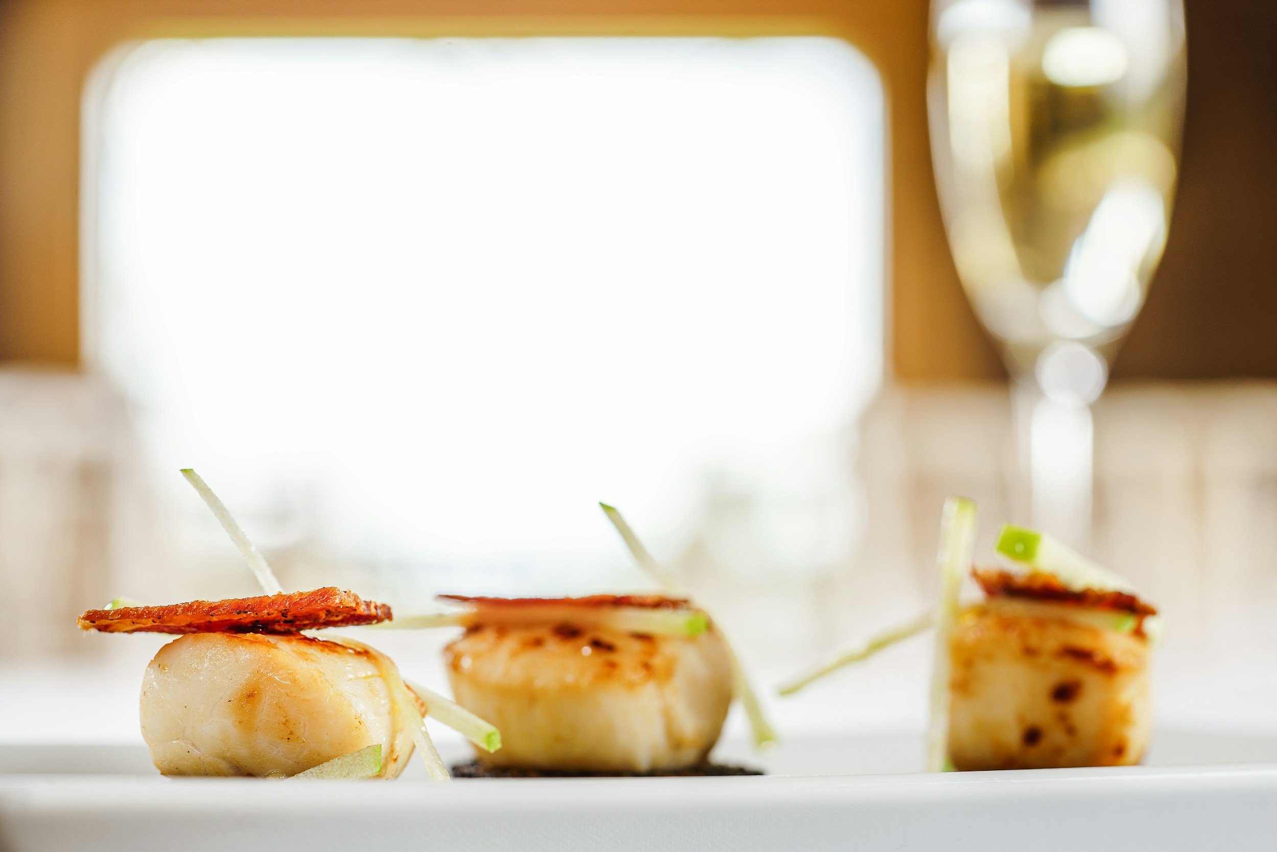 Gourmet seared scallops with crispy topping and garnish, served on a plate with a glass of champagne in the background.