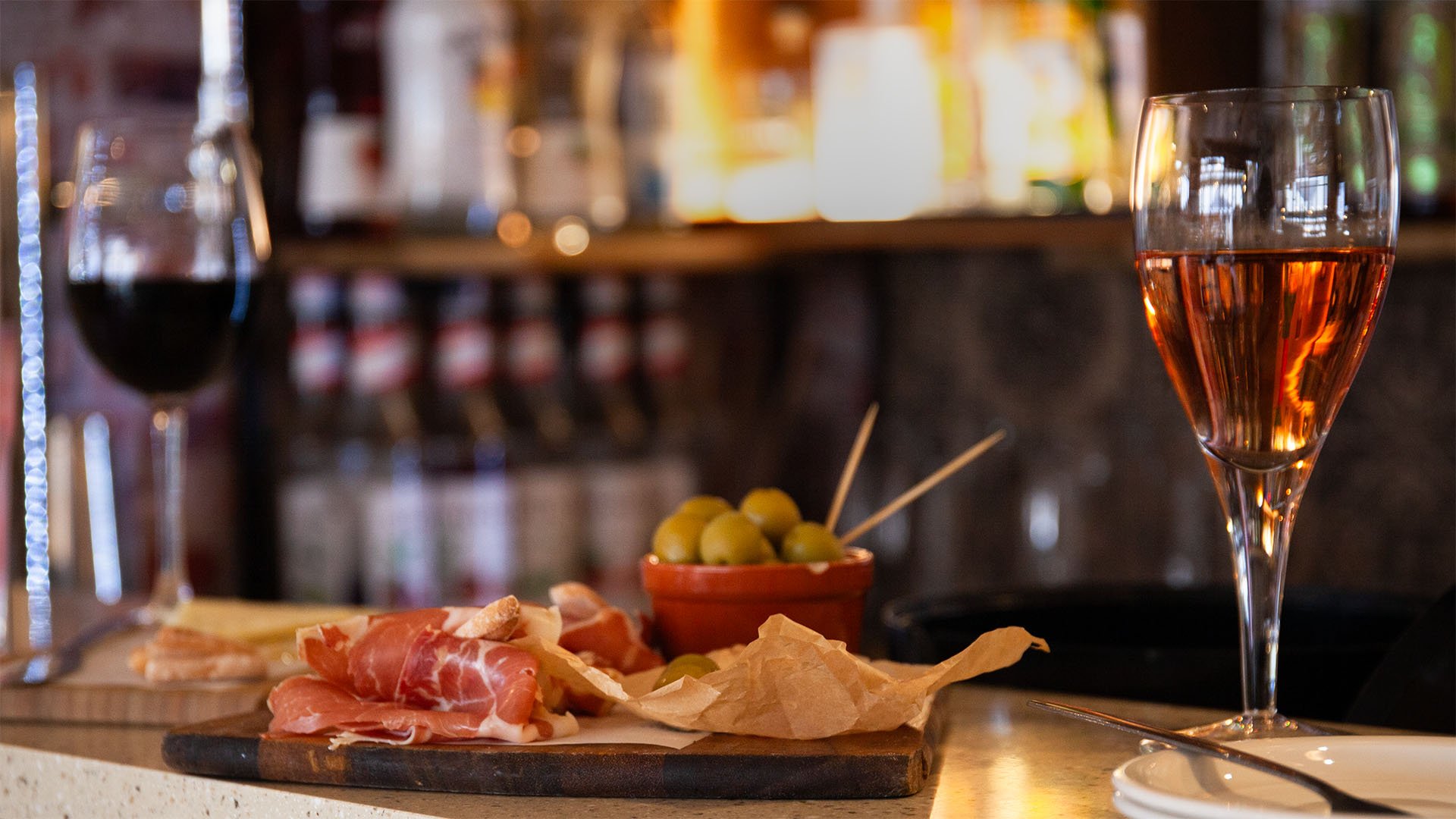 Close-up of charcuterie board with prosciutto, olives, red wine, and rosé in a dimly lit bar setting.