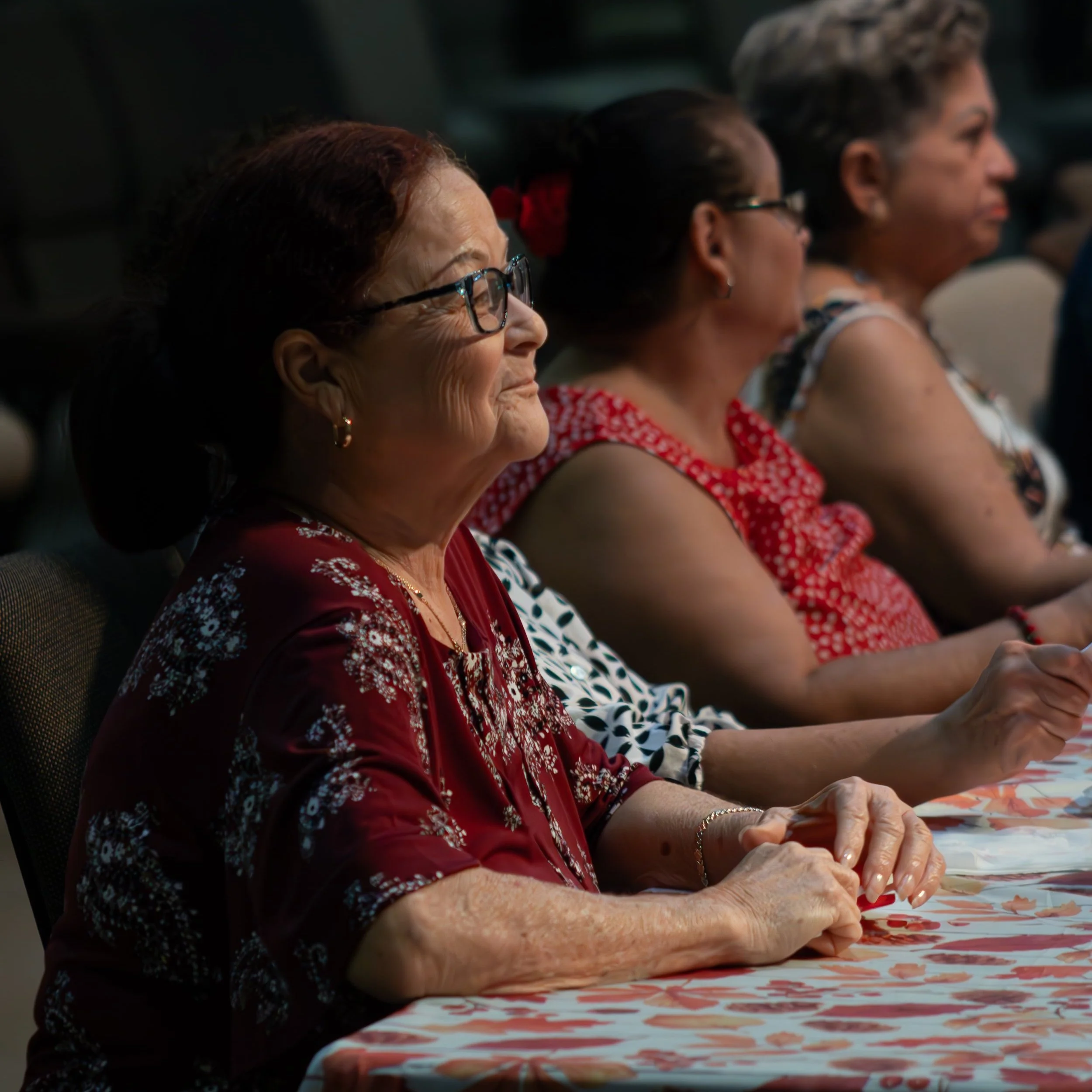 A group of women sitting at a table, attentively listening, with the woman in the foreground wearing glasses and a maroon floral dress.