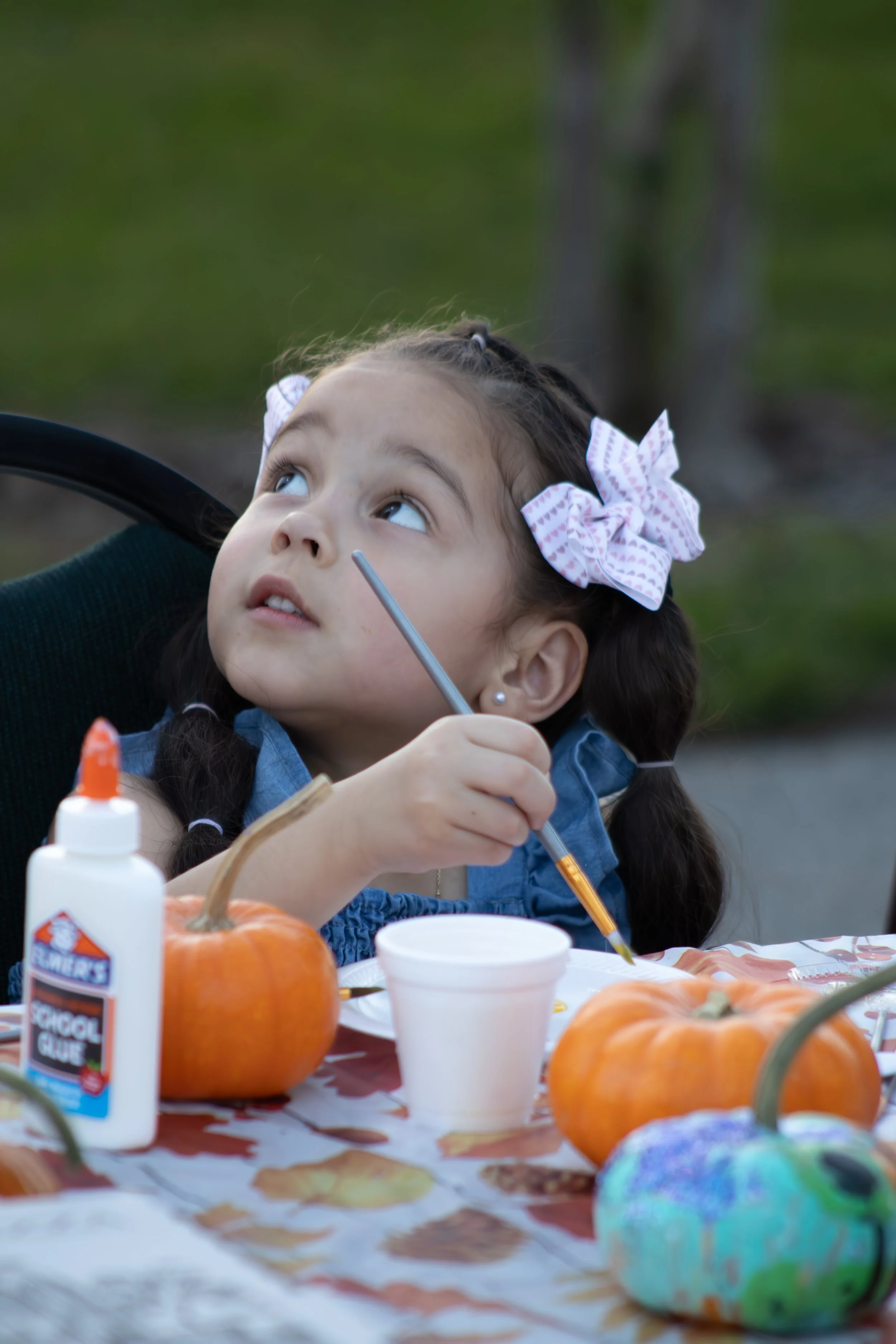Young girl with braided hair and pink bows sitting at an outdoor table surrounded by pumpkins, glue, and paint supplies, holding a paintbrush and looking upward.