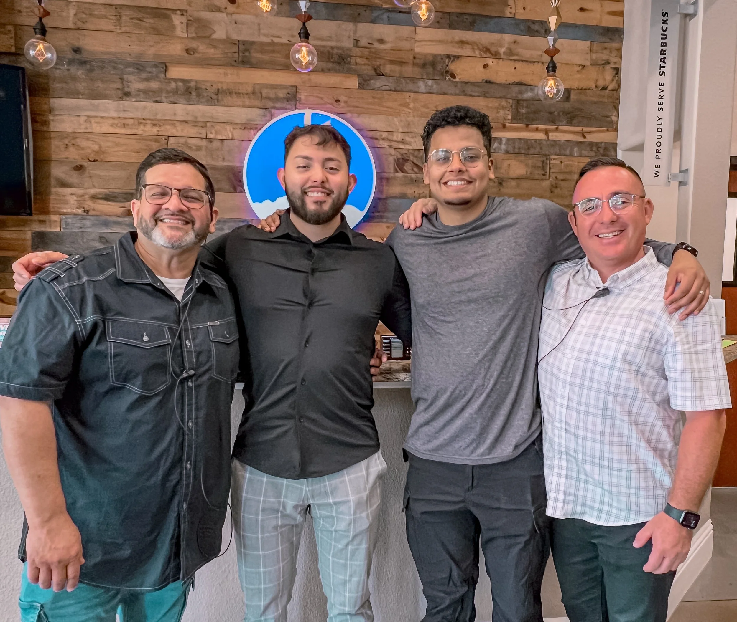 Four men standing together with arms around each other's shoulders inside a restaurant or cafe, smiling at the camera. The background features a wooden wall with a blue circular logo and hanging lightbulbs.