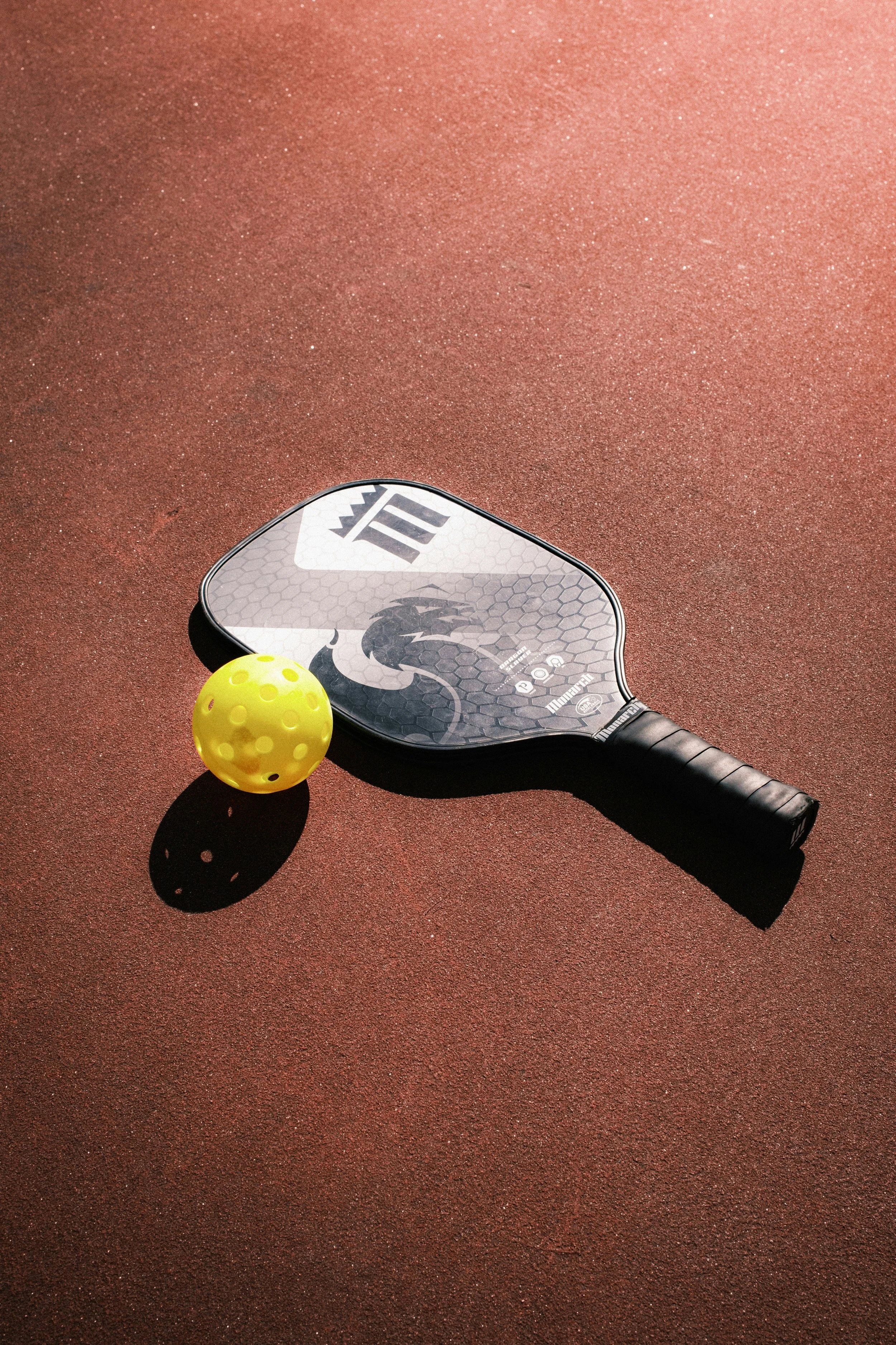 Padel racket and yellow ball on a red court surface.