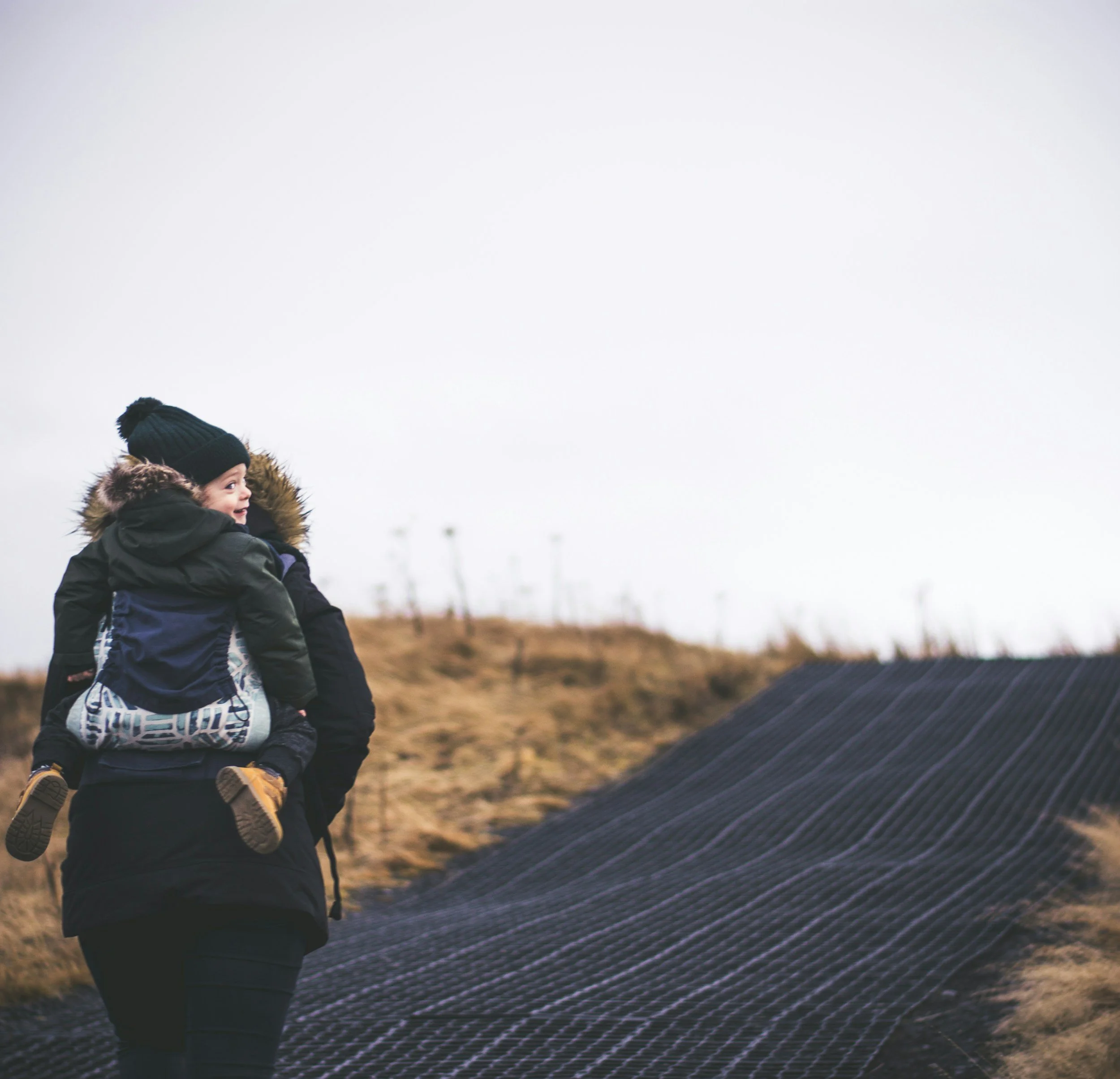 Person carrying a child on their back, both wearing winter clothing, walking on a path surrounded by dry grass.