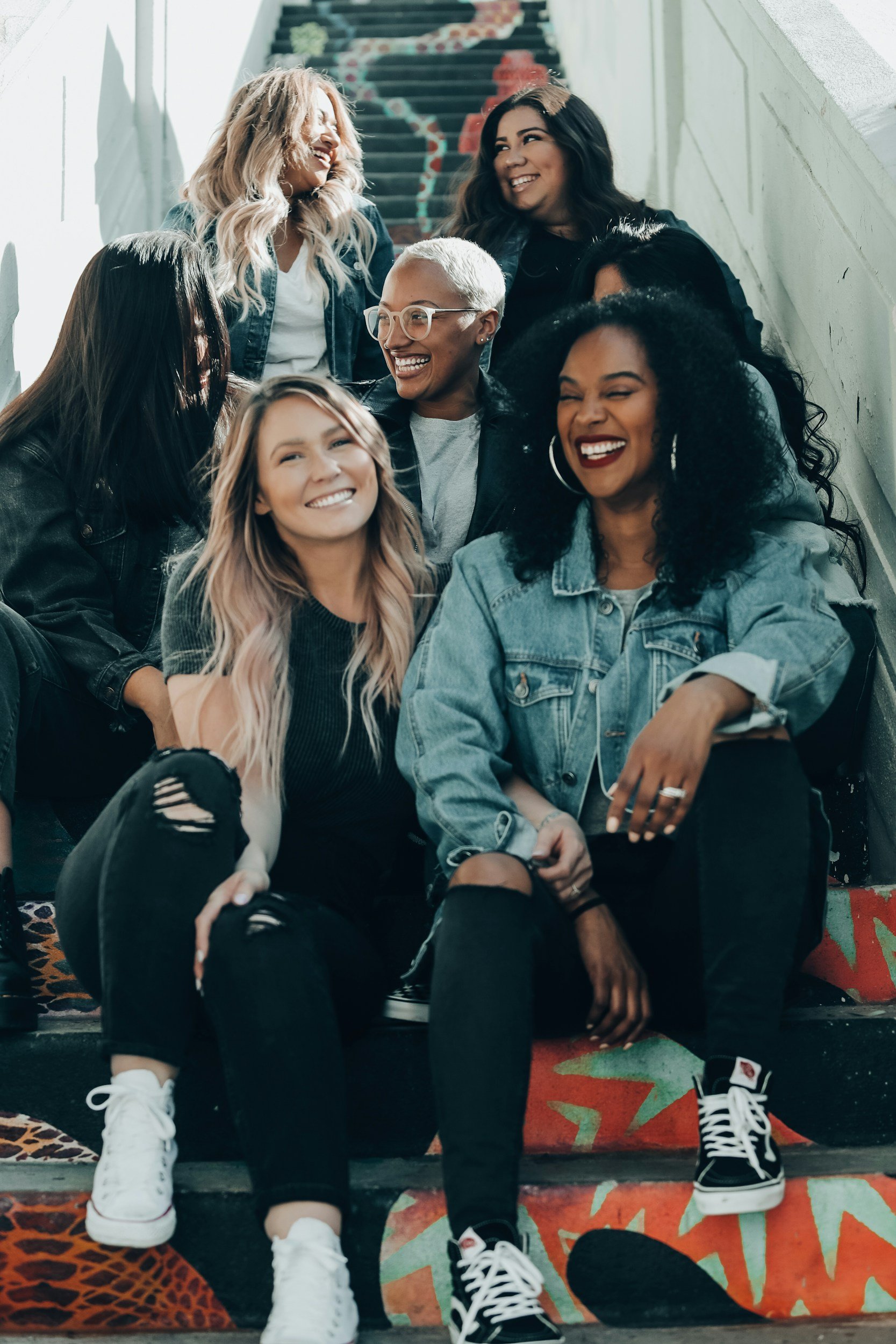 Group of smiling people sitting on colorful stairs, wearing casual clothing like denim jackets and sneakers.
