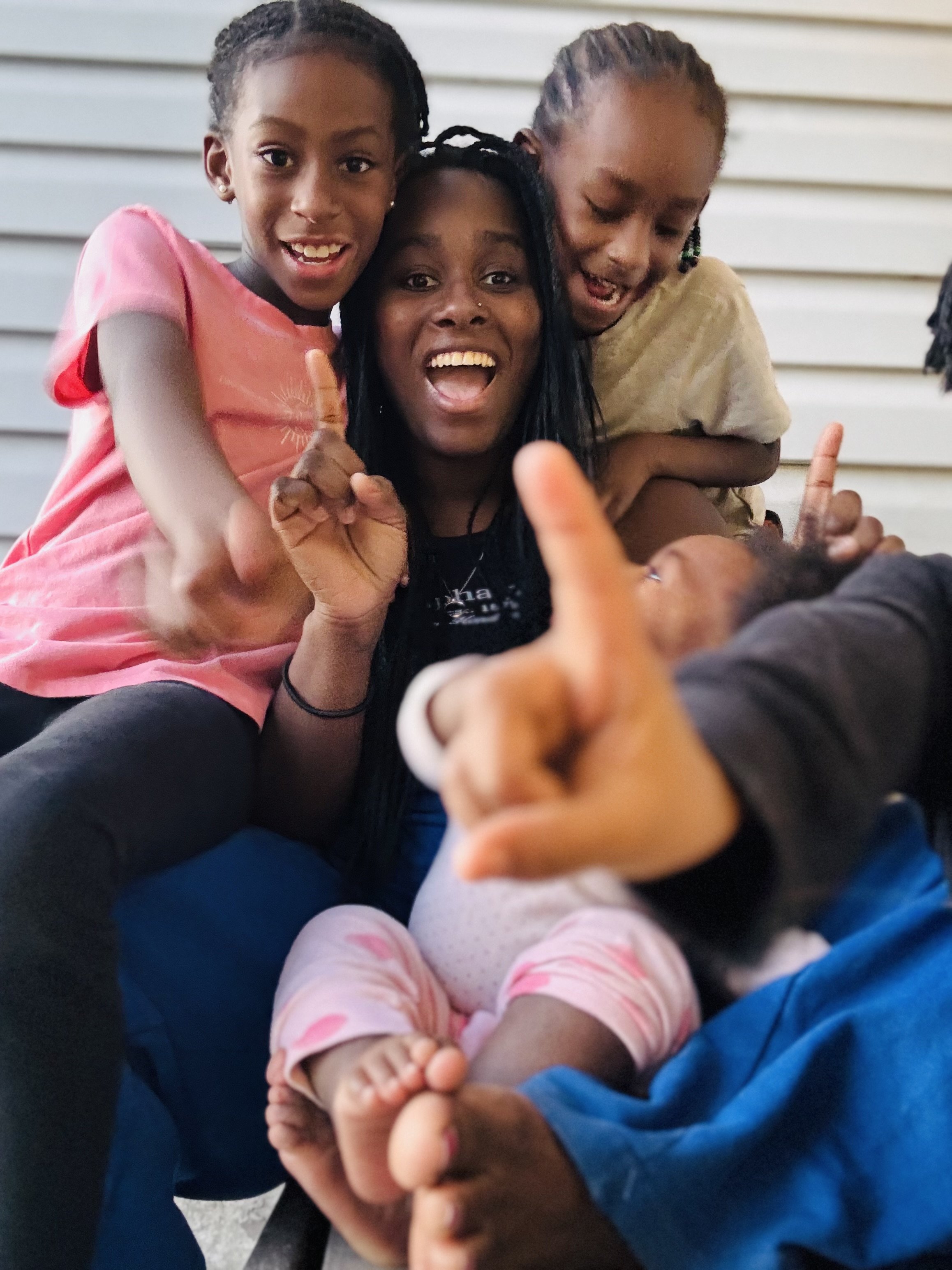 A joyful group of children and an adult, smiling and posing with playful gestures. The adult is central, with one child on their shoulders, and others seated around, showing fingers in fun poses.