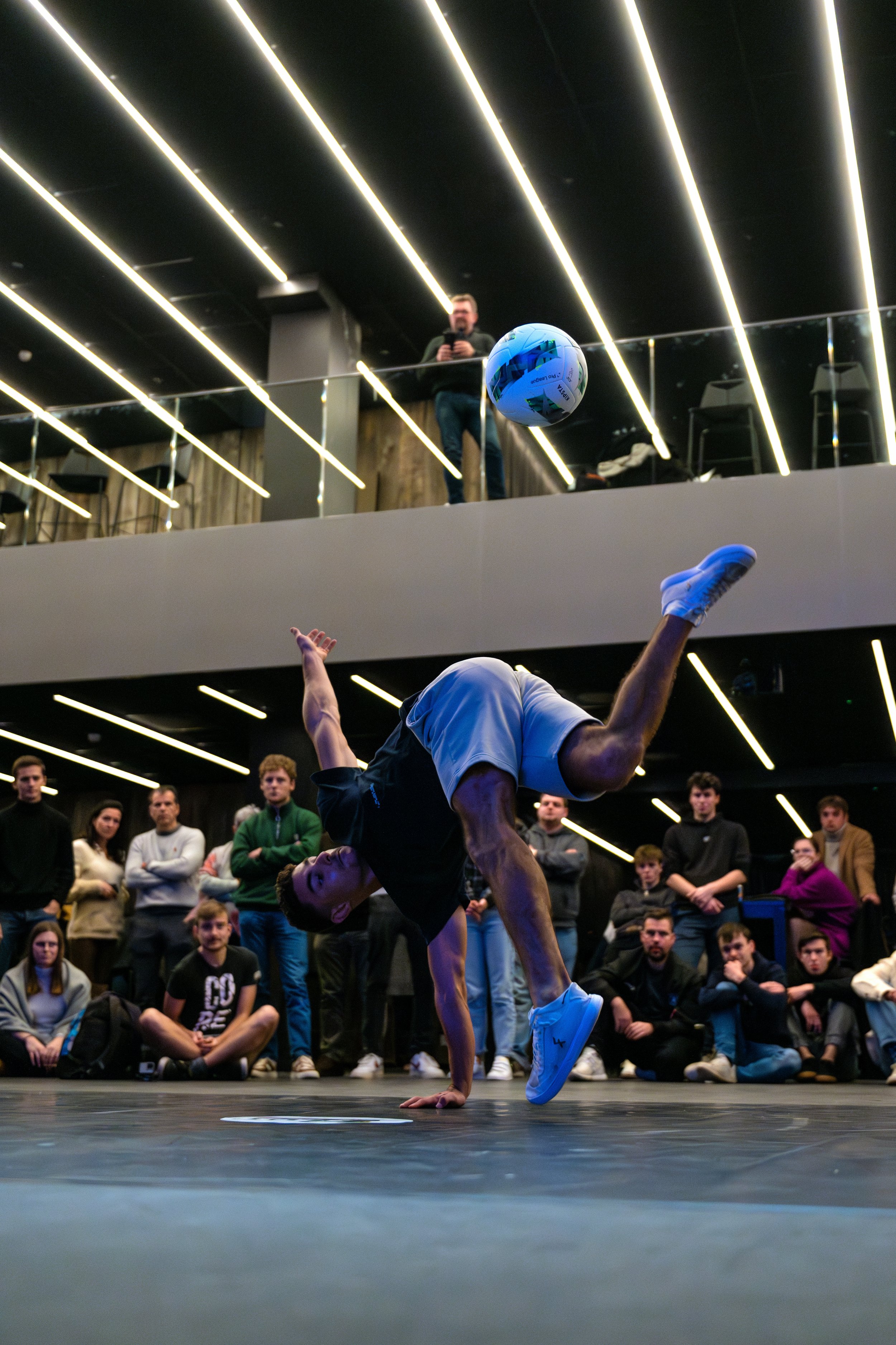 Un danseur breakdance effectue une roue sur un sol noir lors d'une performance, entouré d'un public au fond, avec un plafond noir avec des lumières LED longs et parallèles, et une personne regarde depuis une mezzanine.