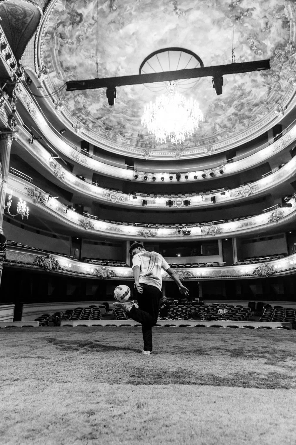 Un homme joue au football dans un théâtre luxueux avec plusieurs étages, des balcons ornés, un plafond peint et un chandelier suspendu.