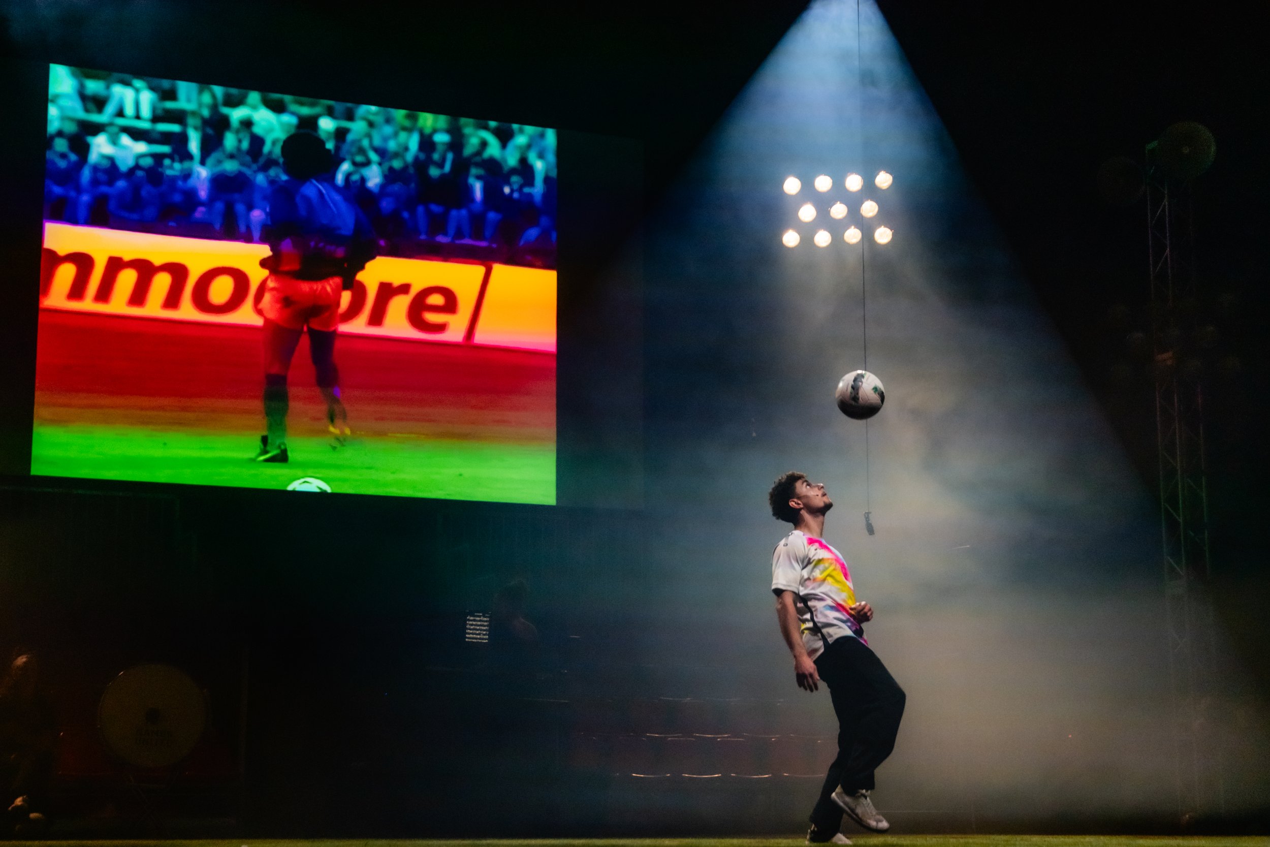 Un homme jongle avec un ballon de football dans une scène sombre, éclairée par un projecteur, avec un grand écran affichant Maradona en arrière-plan.