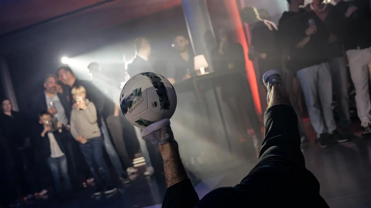 Person lying on the floor with legs in the air holding a soccer ball during a social gathering in a dimly lit room with other people standing and watching.