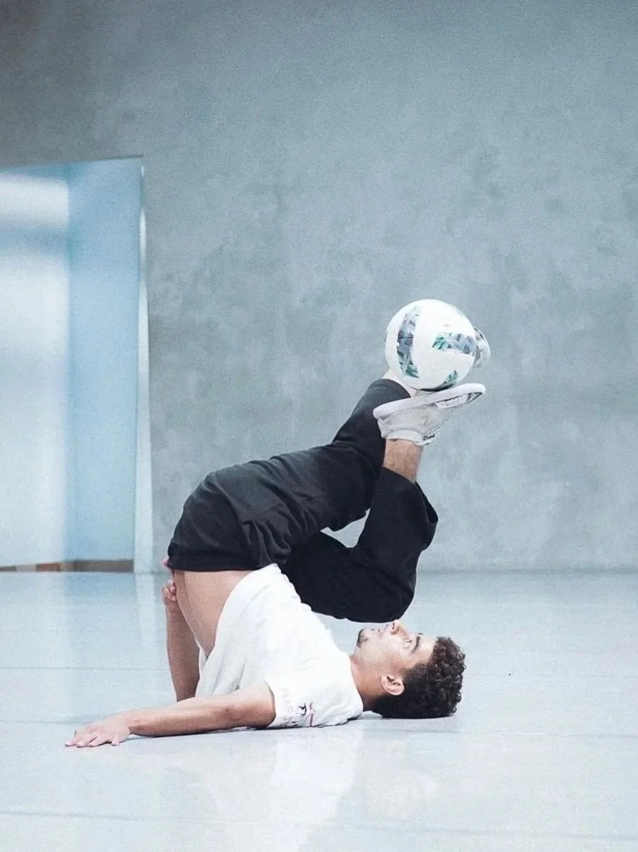 A young man performing a soccer trick on a smooth indoor floor, balancing a soccer ball on his feet while lying on the ground.