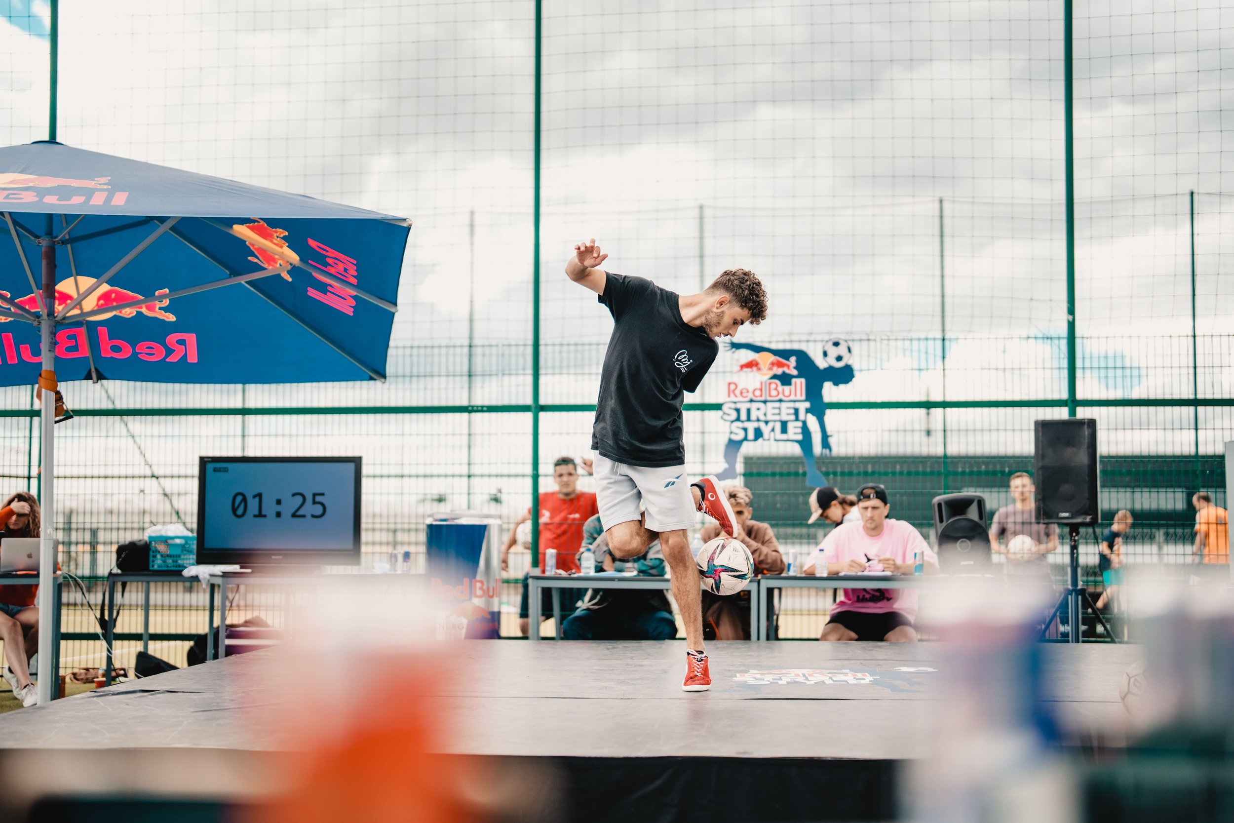 Un jeune homme en t-shirt noir et shorts gris faisant du freestyle avec un ballon de football lors d'une compétition Red Bull Street Style sur une scène extérieure. Des spectateurs et des juges sont derrière une table, avec une horloge de 1:25.