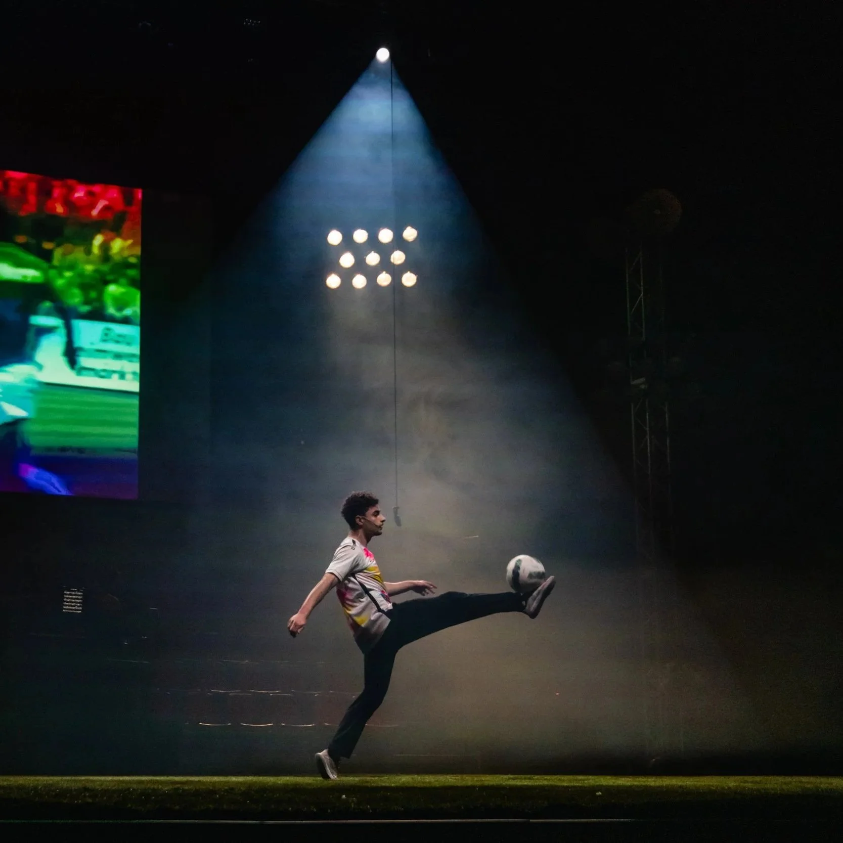 Un garçon joue au football dans une ambiance sombre éclairée par des projecteurs, avec un grand écran lumineux à gauche et un éclairage au plafond.