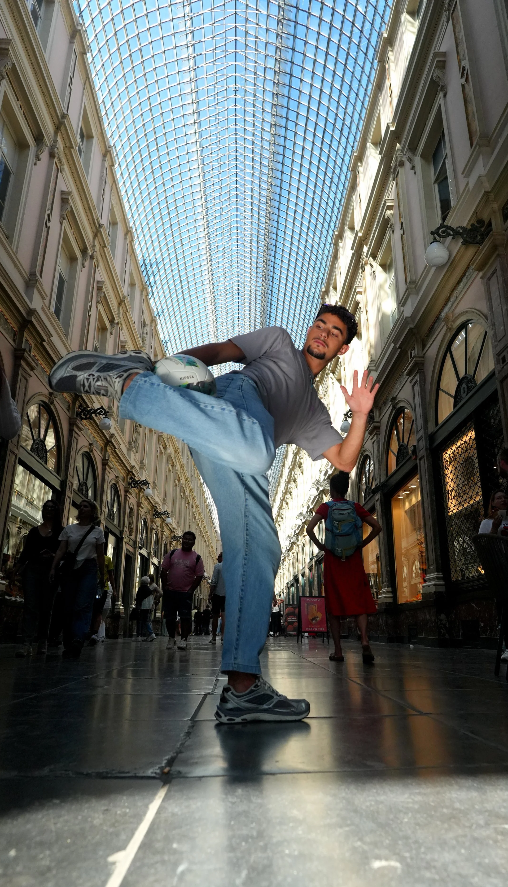 Un homme en jeans et t-shirt gris effectue un saut dans un centre commercial, tenant un ballon de football. Le surplomb de verre au plafond laisse entrer la lumière naturelle, et plusieurs personnes circulent en arrière-plan.