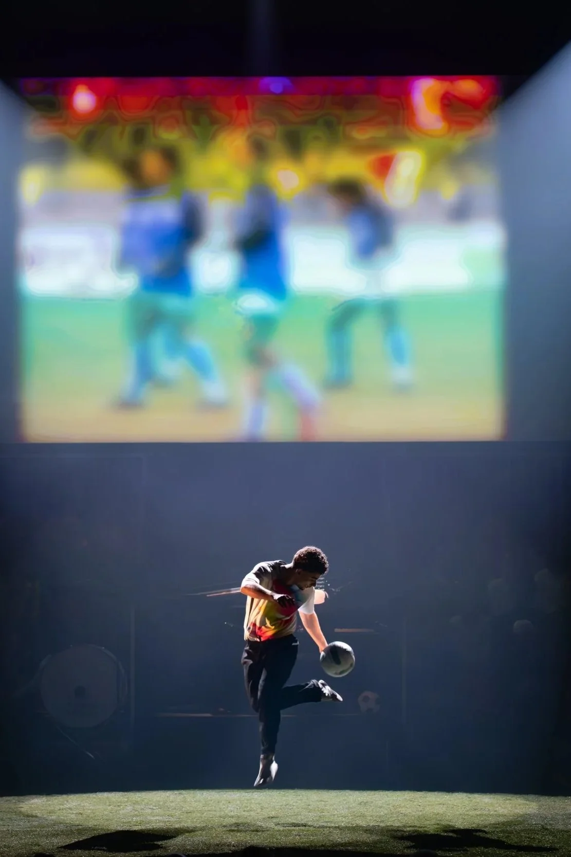 Un jeune homme joue au football sur une scène sombre avec un projecteur, un écran en arrière-plan montre un groupe de personnes dans une autre scène, à l'extérieur.