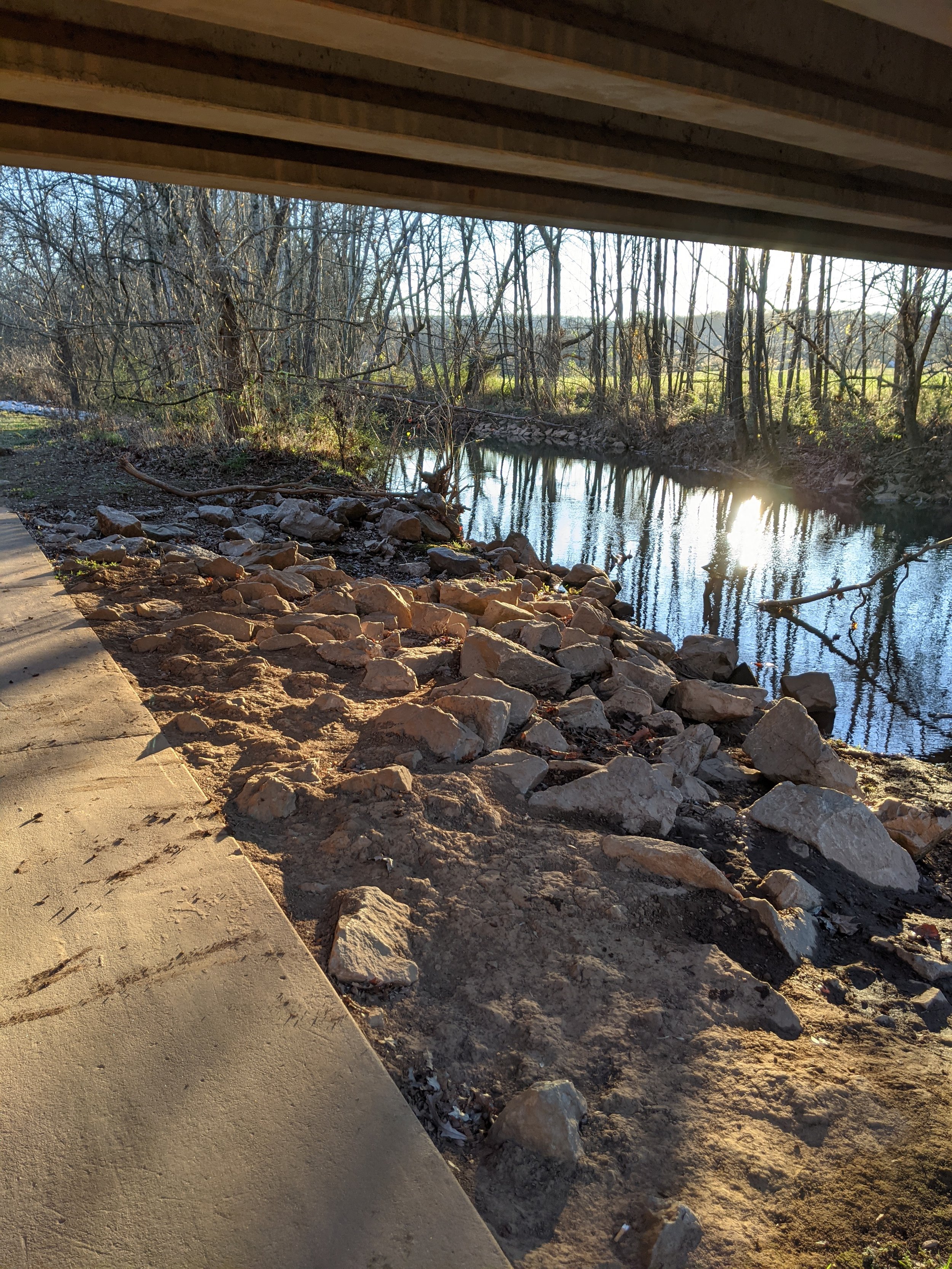 Walkway lined with rocks next to a creek, passing under a bridge at sunset.
