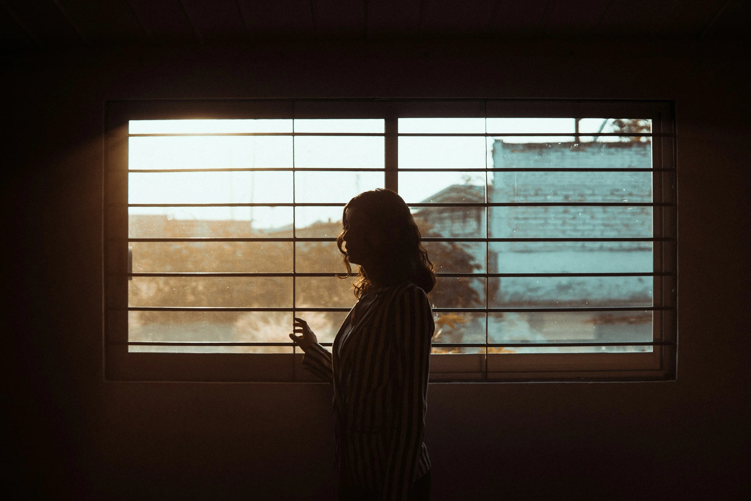 Silhouette of a woman standing near a window with horizontal bars, with a view of buildings and trees outside during sunset or sunrise.