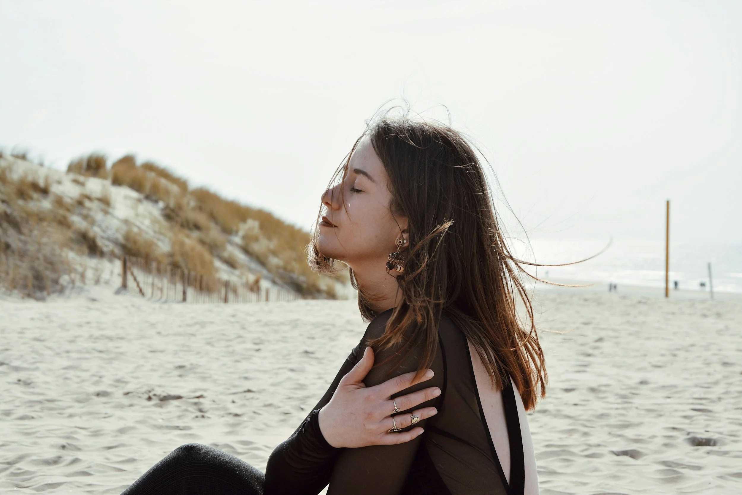 Young woman with brown hair and earrings sitting on a beach with eyes closed, wearing black clothing, and holding her shoulder, with sand dunes and volleyball poles in the background.