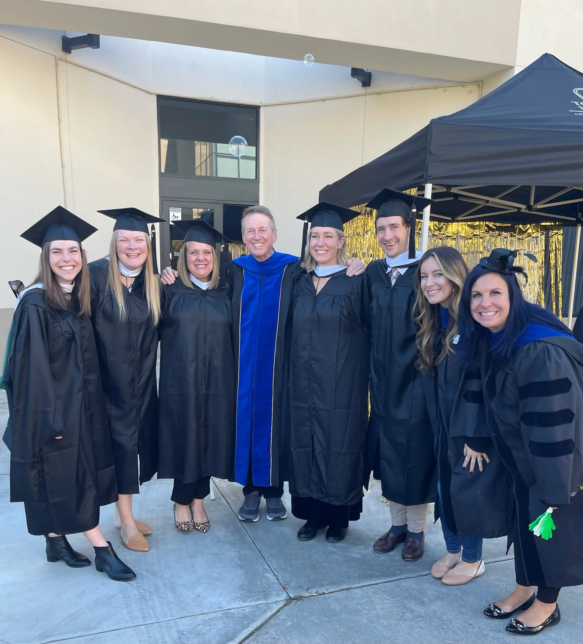 Group of seven people in graduation gowns and caps posing with a man in academic regalia at outdoor graduation ceremony.