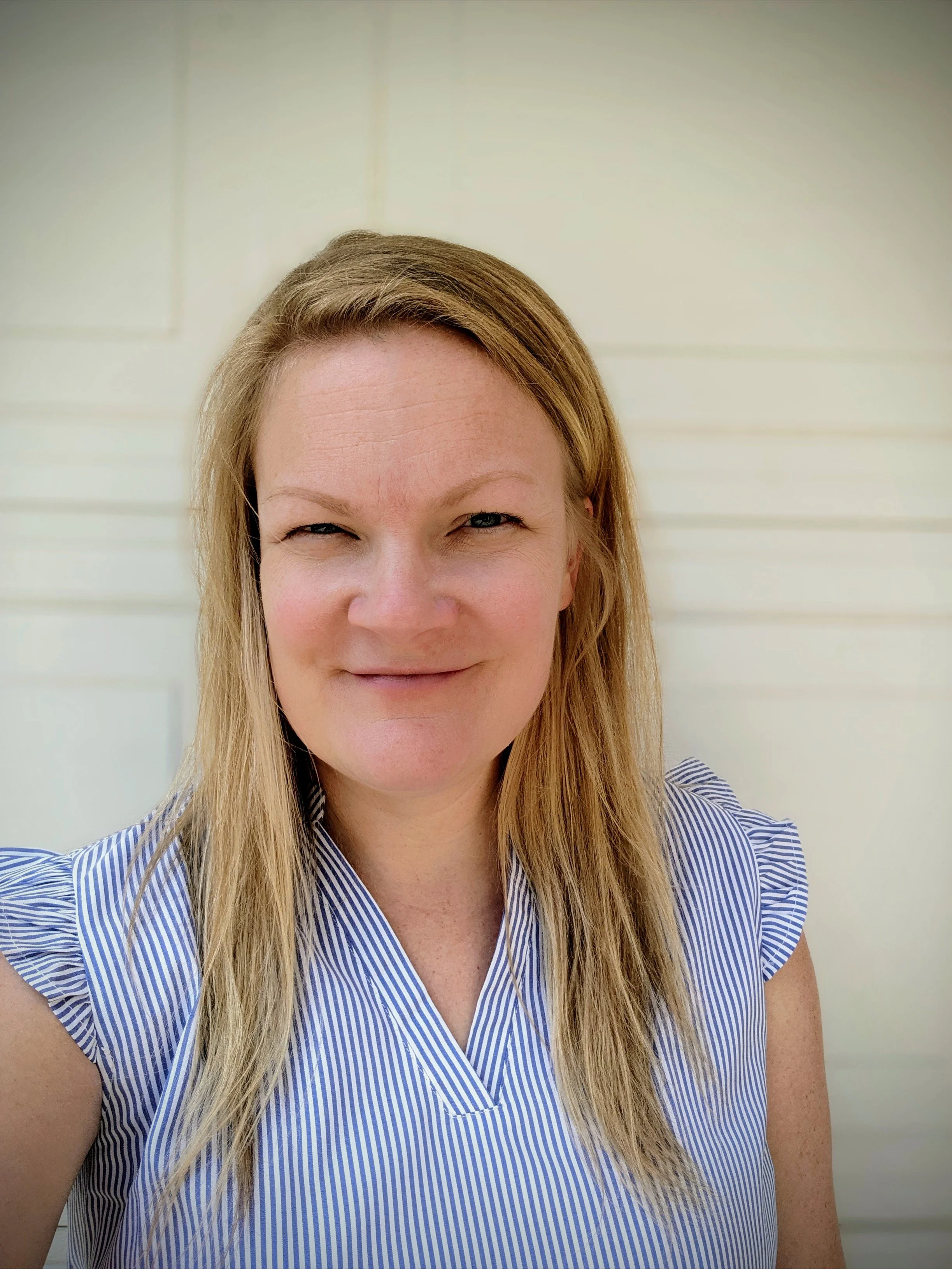A woman with light hair smiling outdoors, wearing a blue and white striped top, standing in front of a light-colored wall.