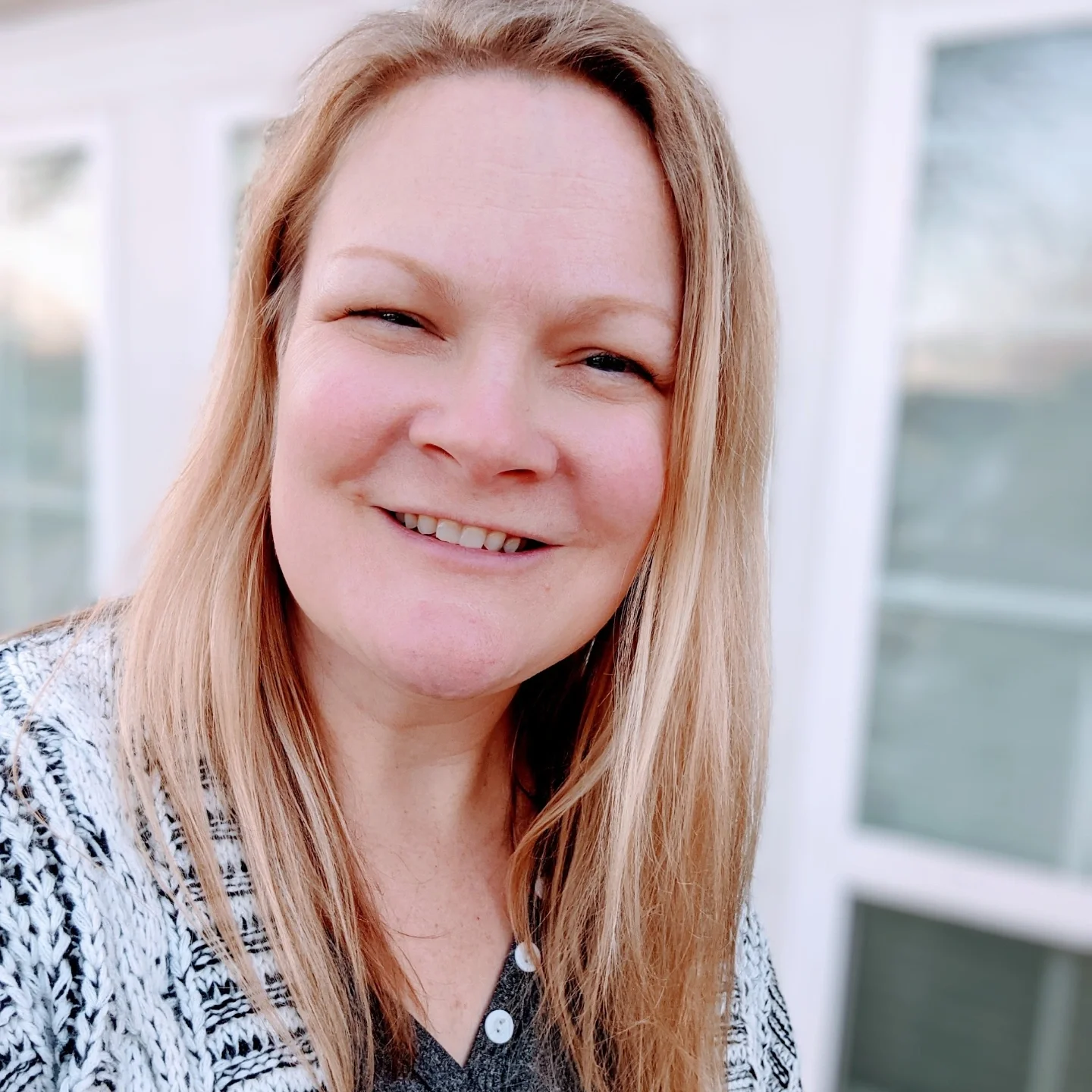 A woman with long reddish-blond hair smiling outdoors with windows in the background.