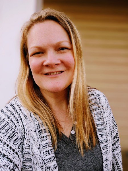 A woman with long blonde hair smiling, wearing a patterned cardigan over a dark top, standing indoors with a wooden background.