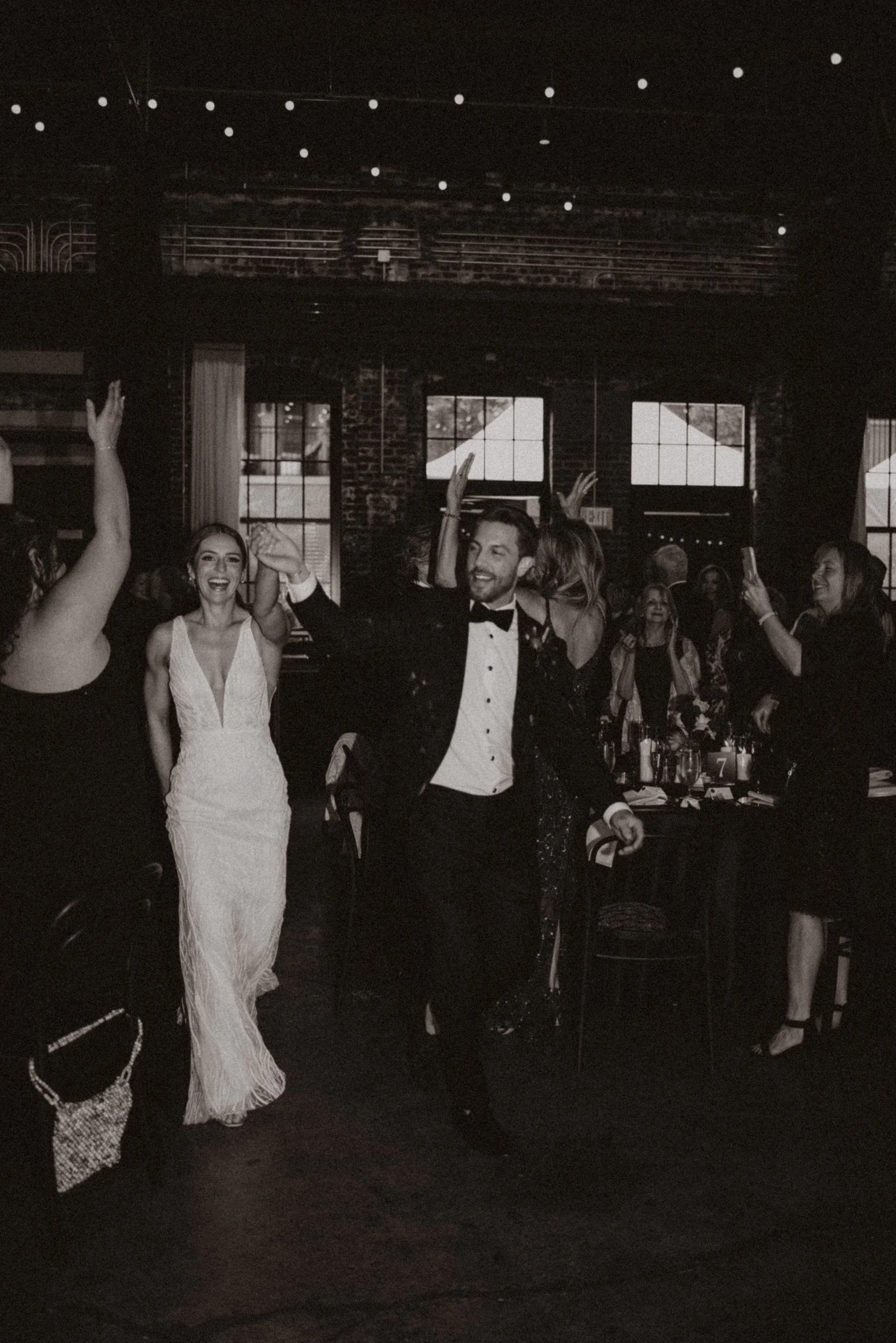 People dancing and celebrating at a party or event in a dimly lit room with large windows and exposed brick walls.