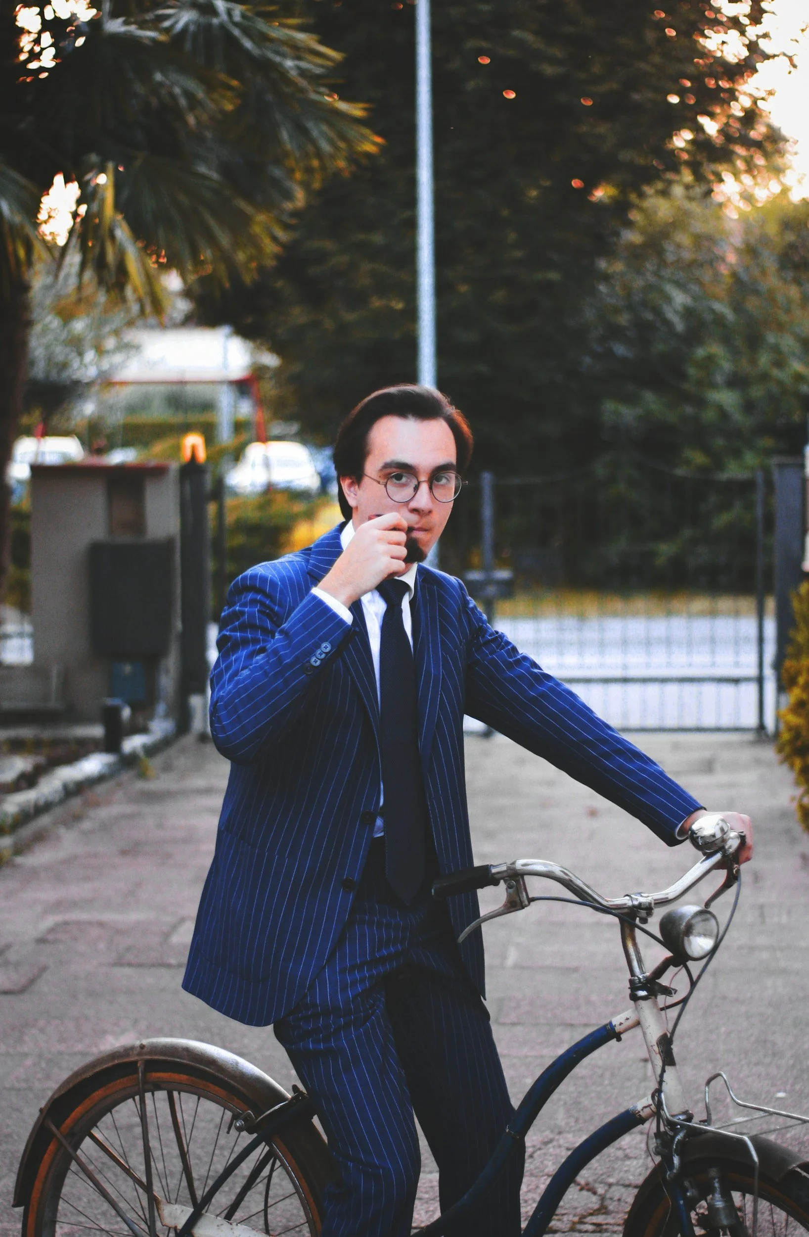 A man in a blue pinstripe suit and glasses is sitting on a bicycle outdoors in a park at sunset.