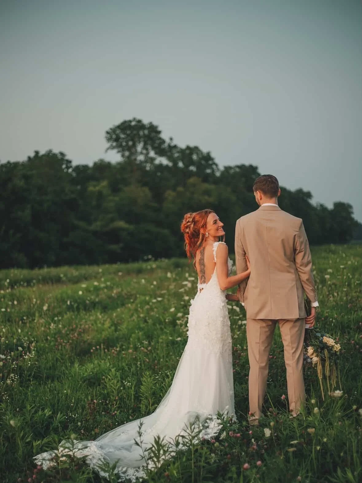 A bride and groom standing in a field at sunset, holding hands and smiling at each other.