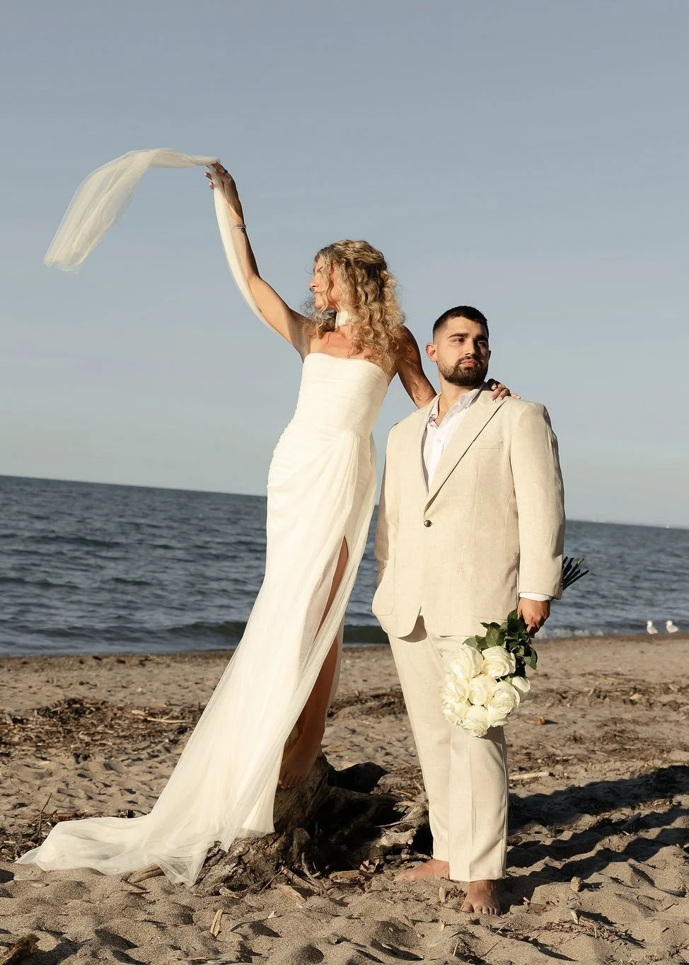 A couple dressed in wedding attire standing on a sandy beach at sunset, with the woman on a piece of driftwood holding a sheer cloth in the air, and the man holding a bouquet of white roses.
