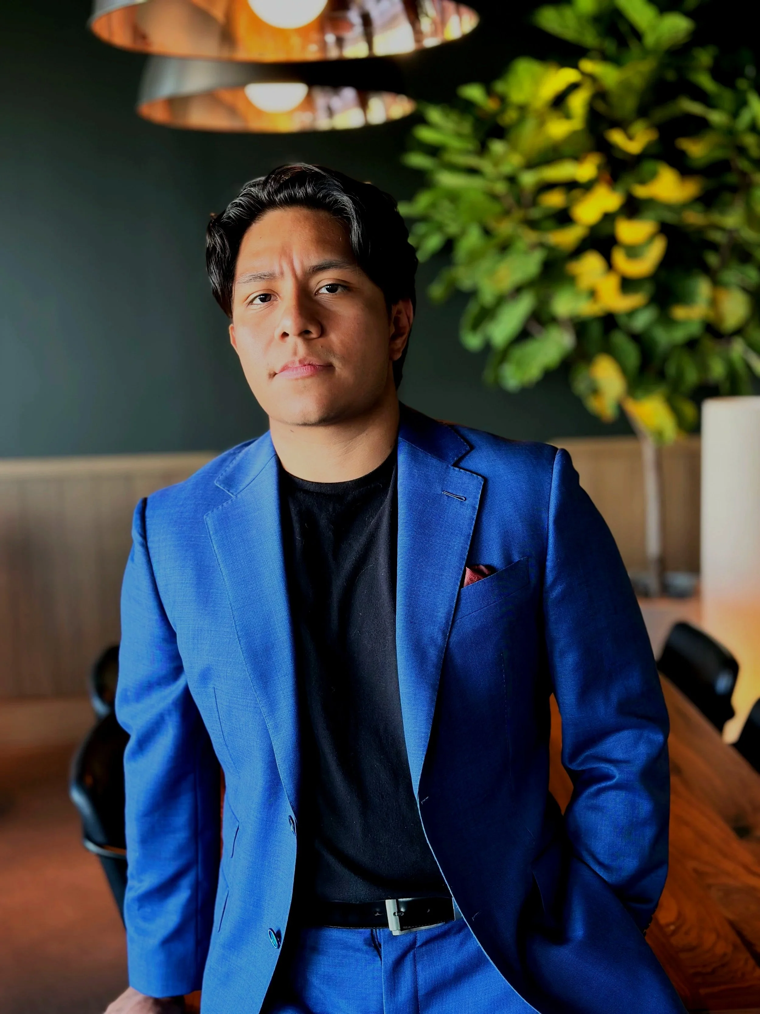 A man in a blue suit with a black shirt sitting in a room with a large green plant and copper hanging lamps.