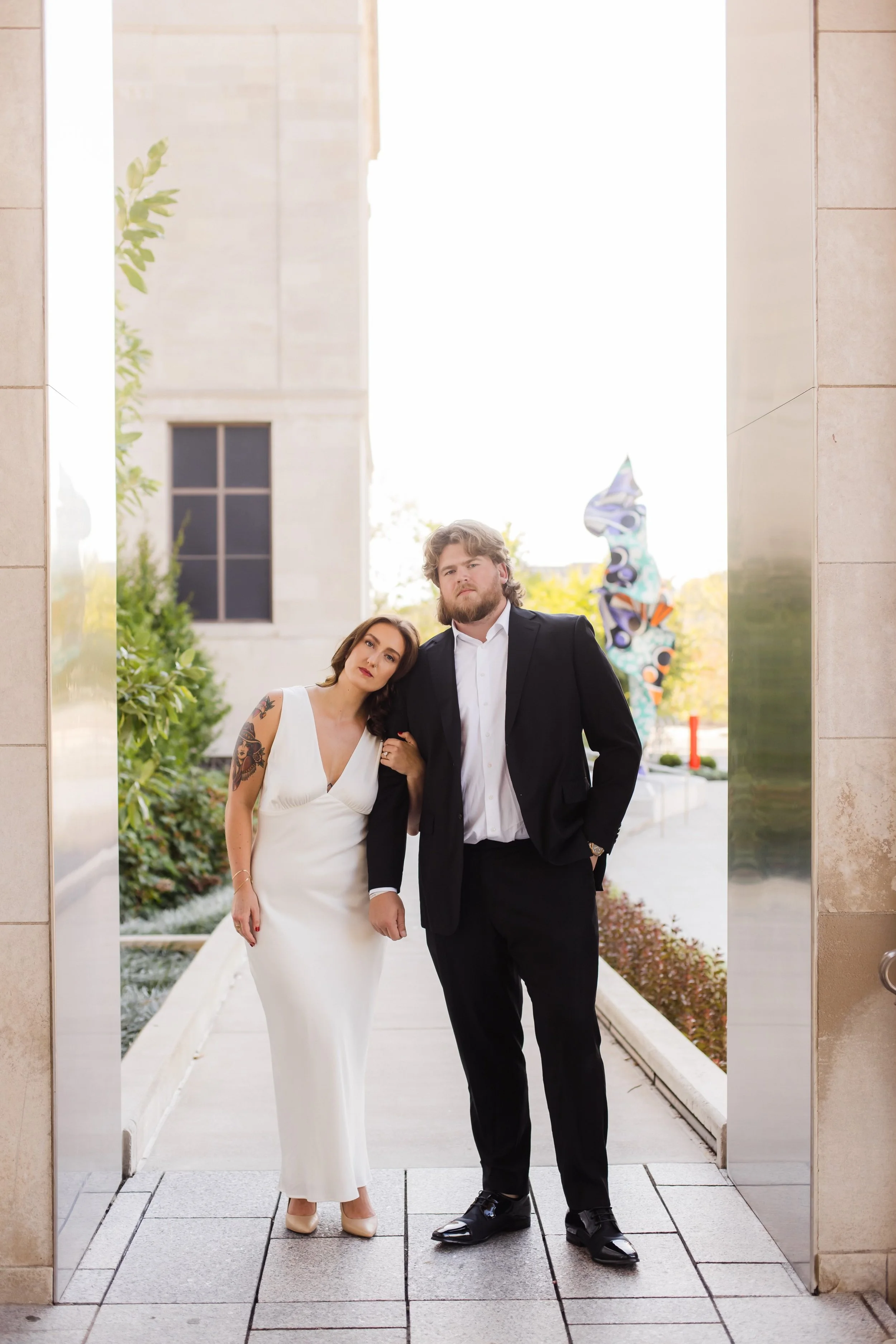 A man and a woman standing close together in wedding attire, outdoors, under an archway with trees and a colorful sculpture in the background.