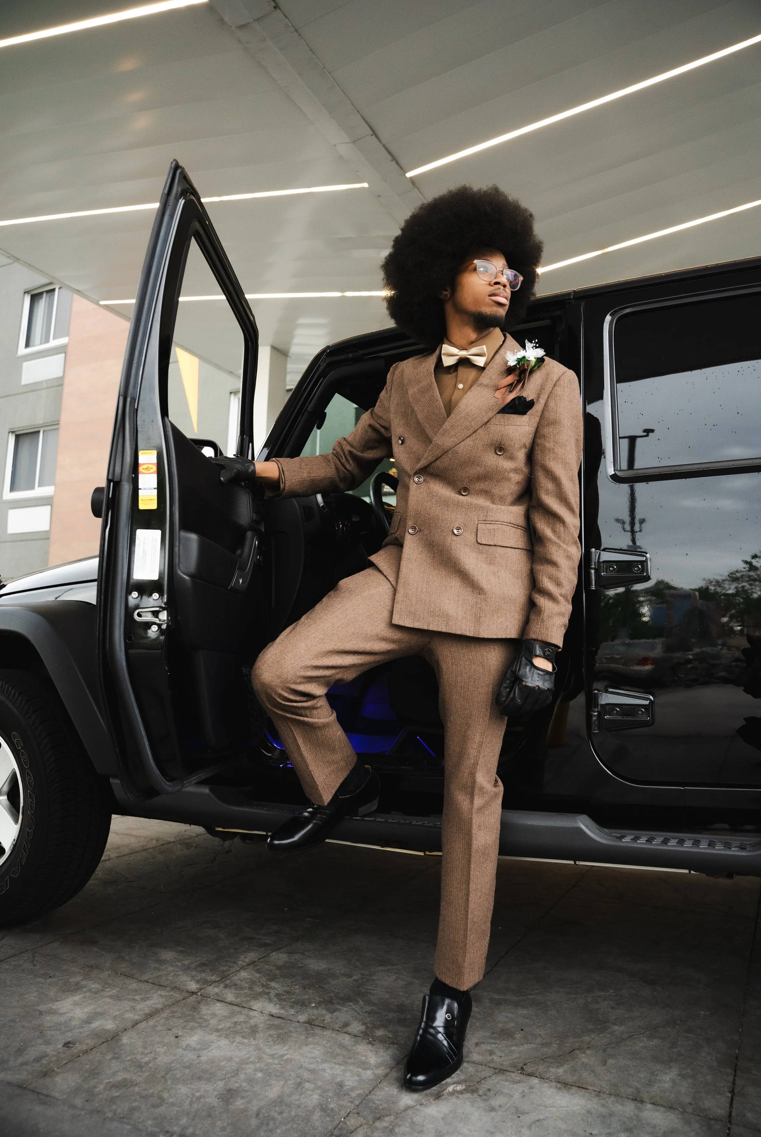 A man with an afro hairstyle, glasses, and a mustache, dressed in a brown double-breasted suit and black boots, leaning against a black vehicle and looking away.