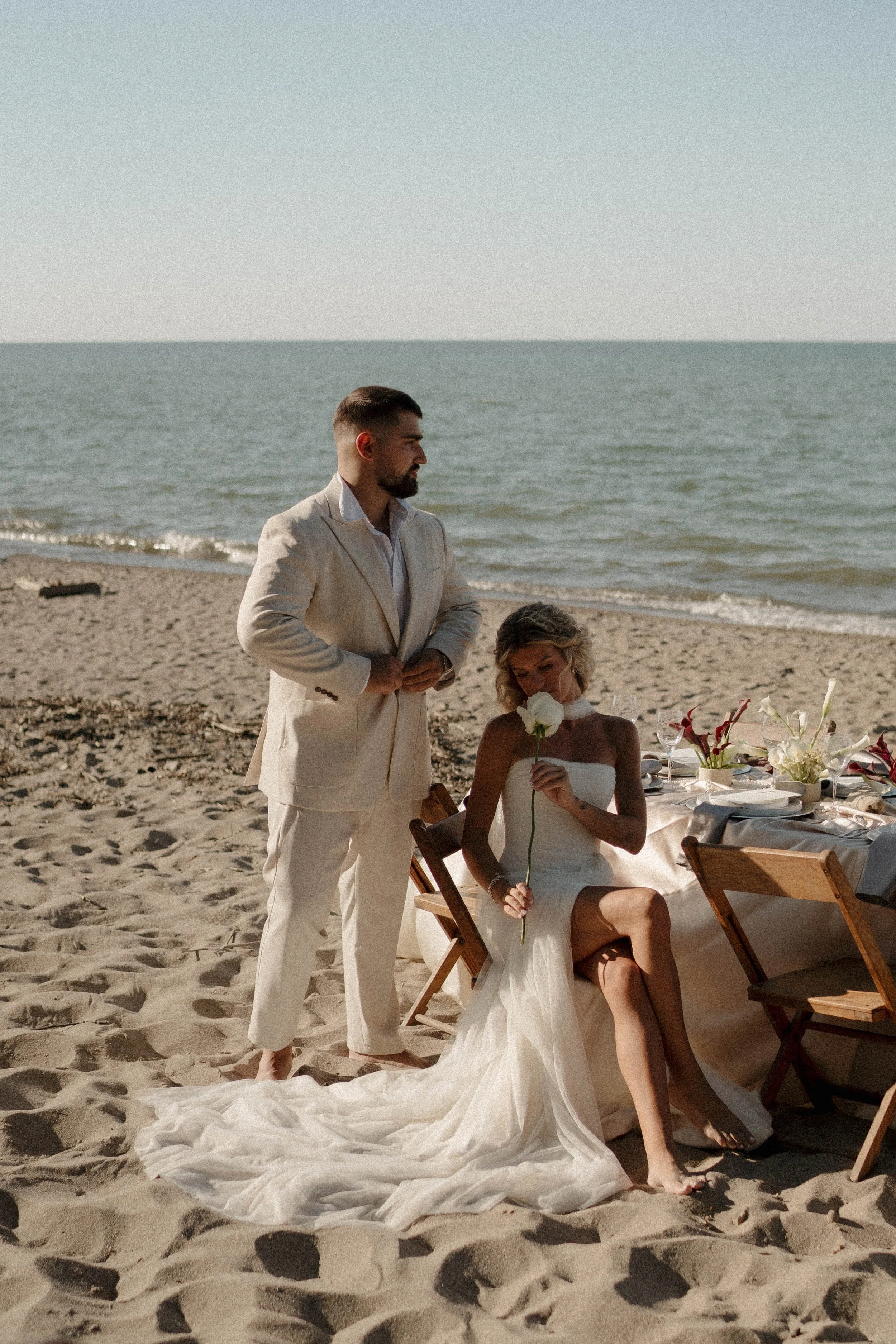 A man and a woman in wedding attire on a sandy beach with the ocean in the background. The woman is seated and holding a white flower, while the man stands beside her, dressed in a beige suit.