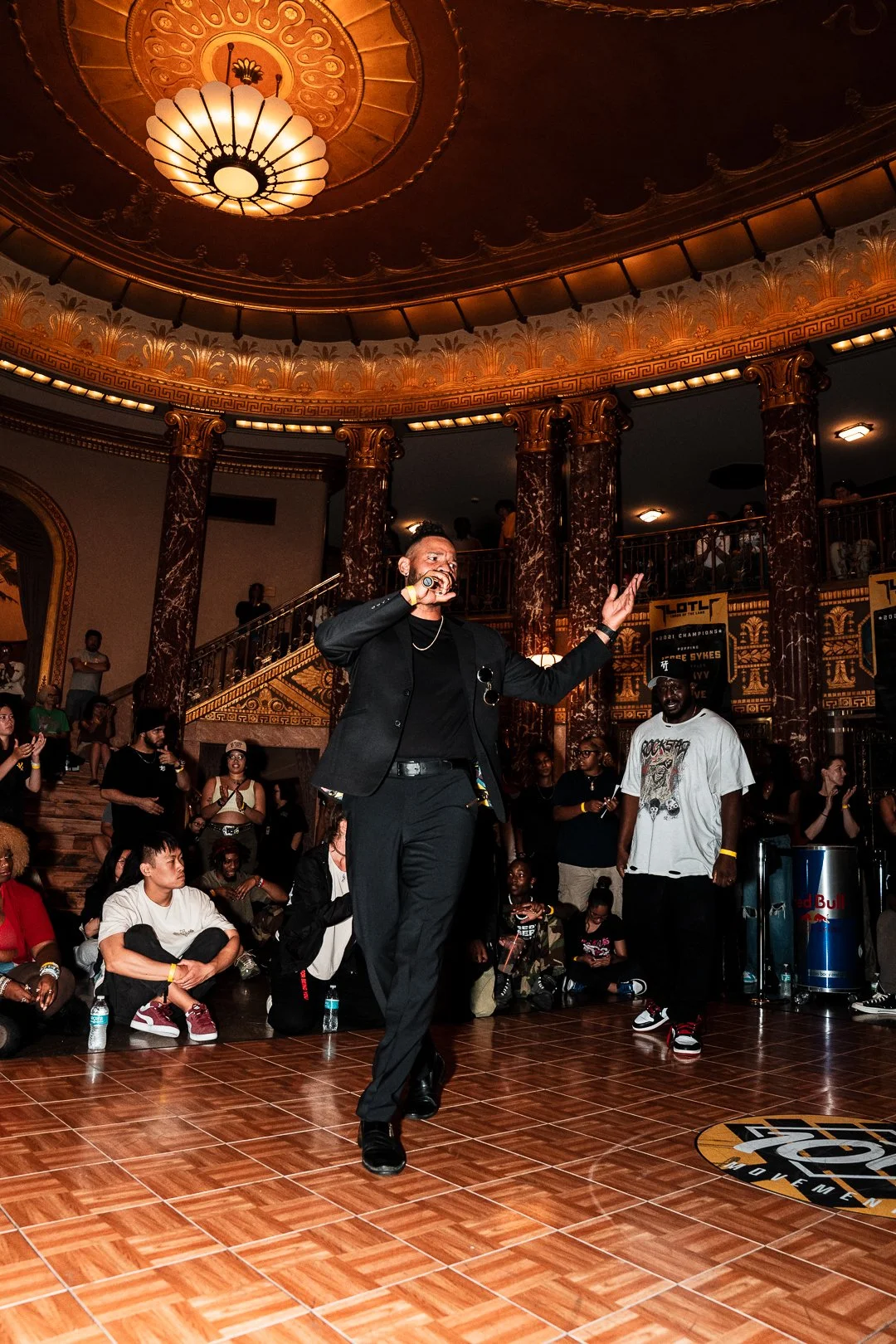 A man in black suit and black shirt singing into a microphone on a wooden floor in an ornate, grand hall with gold and dark red decor. Audience members sit and stand around watching, some with water bottles, and a large Red Bull cooler is visible to 