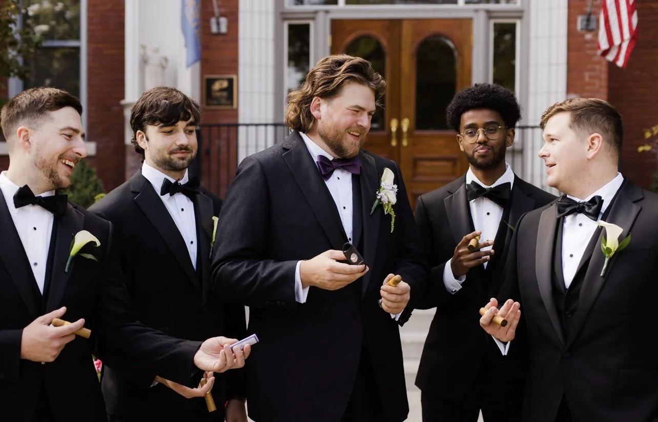 A group of men in tuxedos with boutonnieres, gathered outside a building with wood double doors. They are smiling and holding cigars, sharing a moment of celebration.
