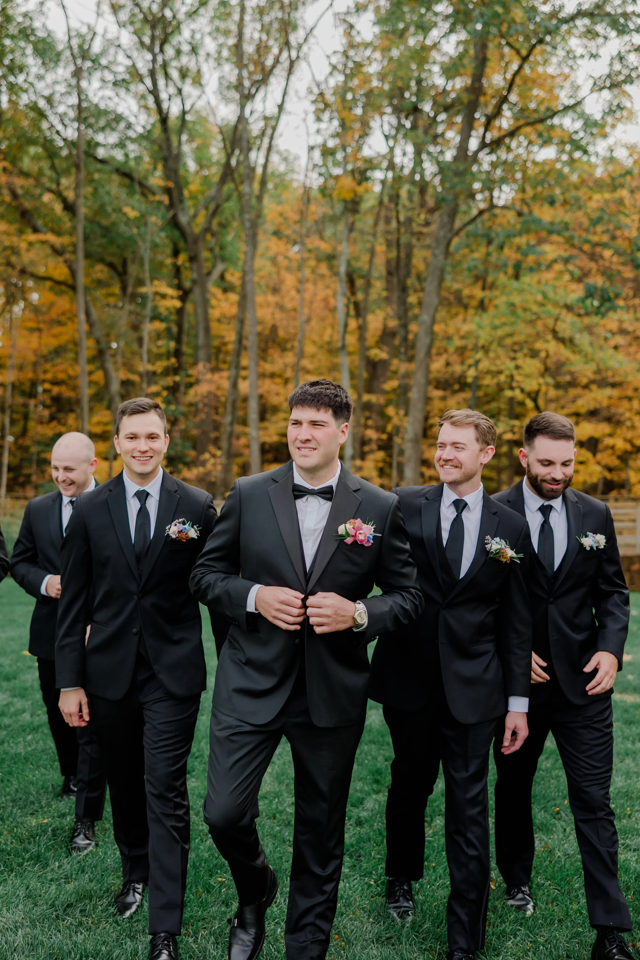 A group of men in black tuxedos walking on a grassy field with fall-colored trees in the background.