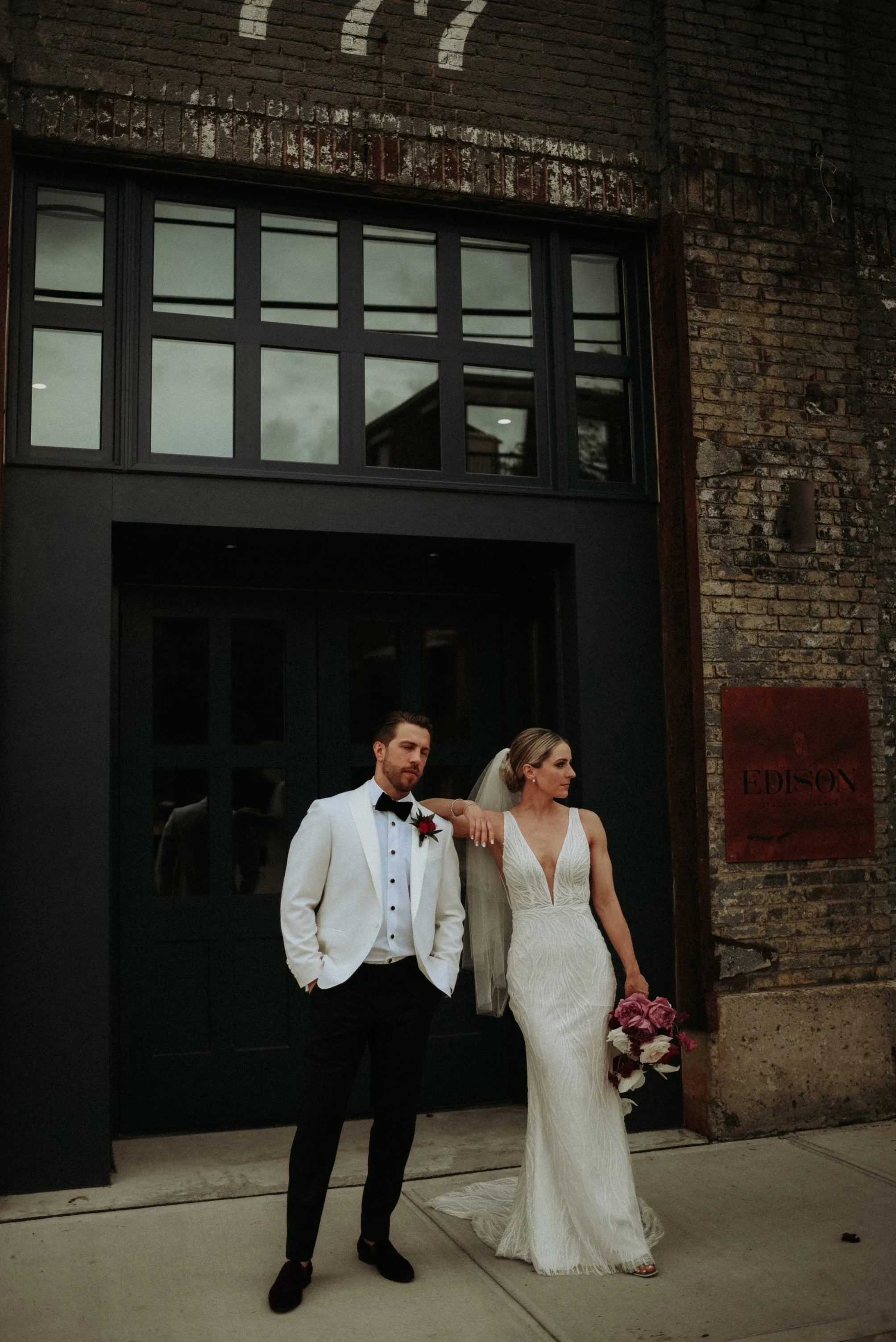 A bride and groom standing outside a brick building, dressed in wedding attire, with the groom in a white tuxedo jacket and the bride in a white lace gown holding a bouquet of flowers.