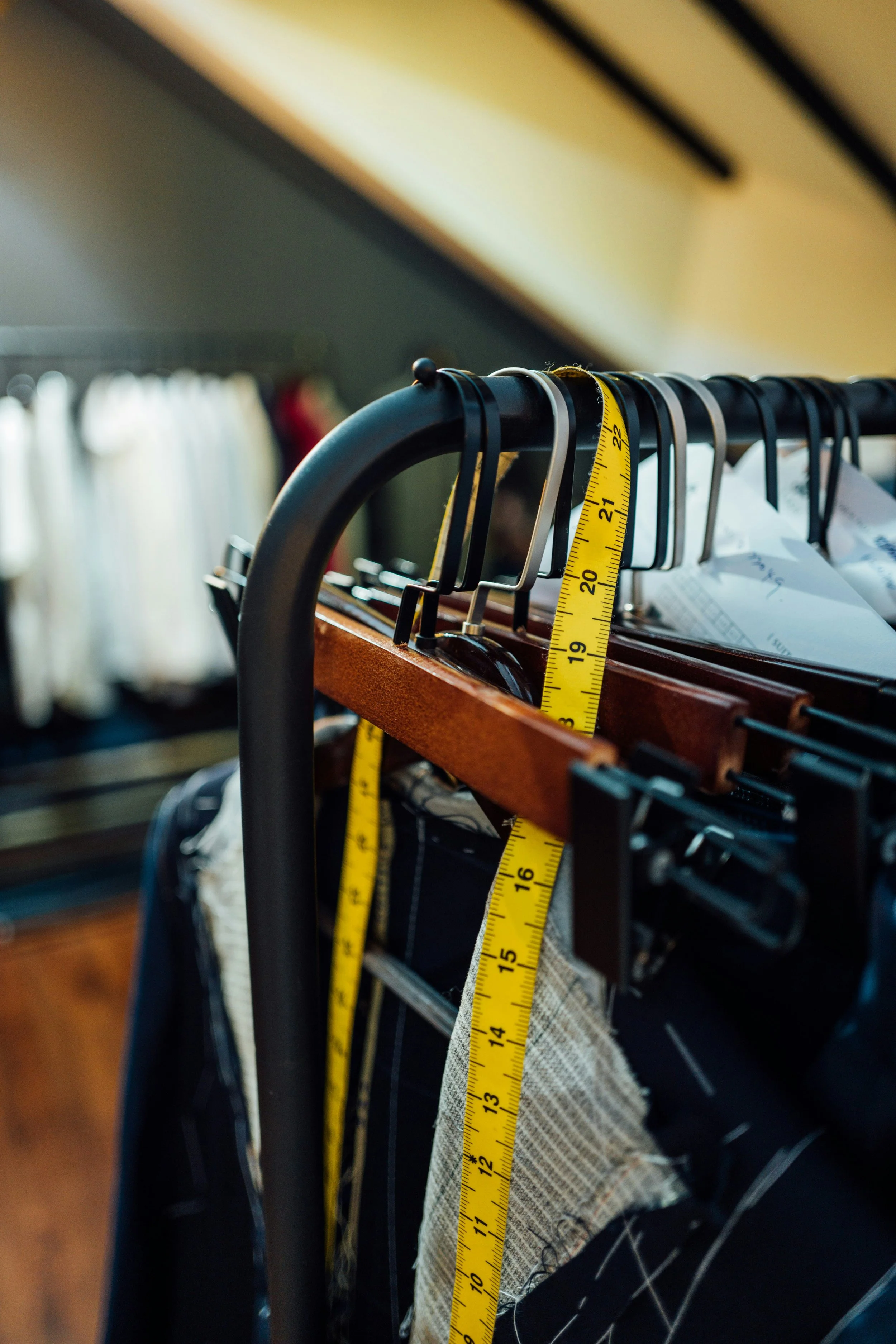 Clothing rack with various shirts, pants, and fabrics hanging on black metal hangers, measuring tape draped over the rack, in a room with wooden flooring and a window with white curtains.