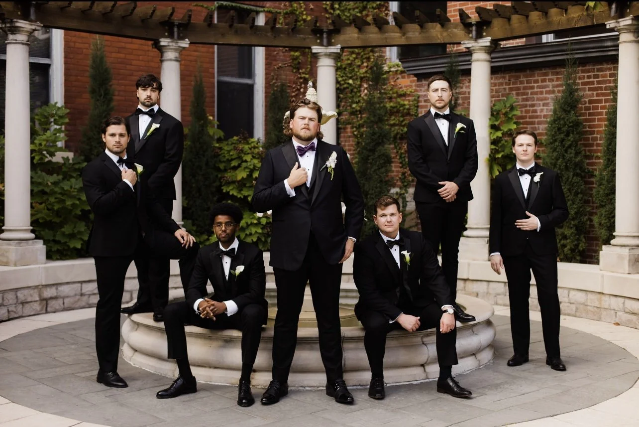 A group of eight men dressed in black tuxedos with bow ties, posing around a fountain in an outdoor garden setting for a formal photo.