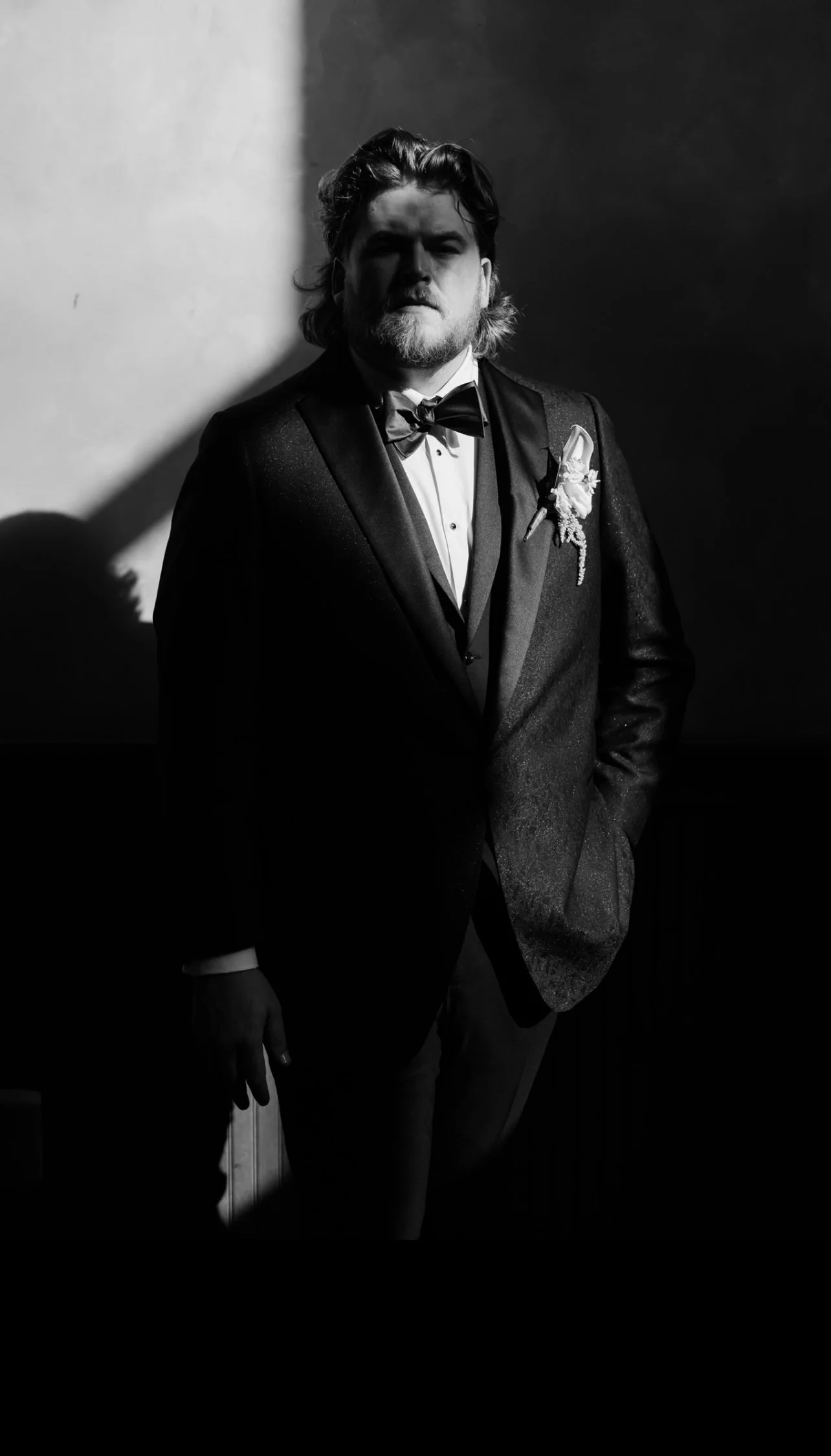 A man with a beard and long hair dressed in a tuxedo with a bow tie. He is standing in a dark room with a light source on his left, creating a dramatic shadow on the wall beside him.